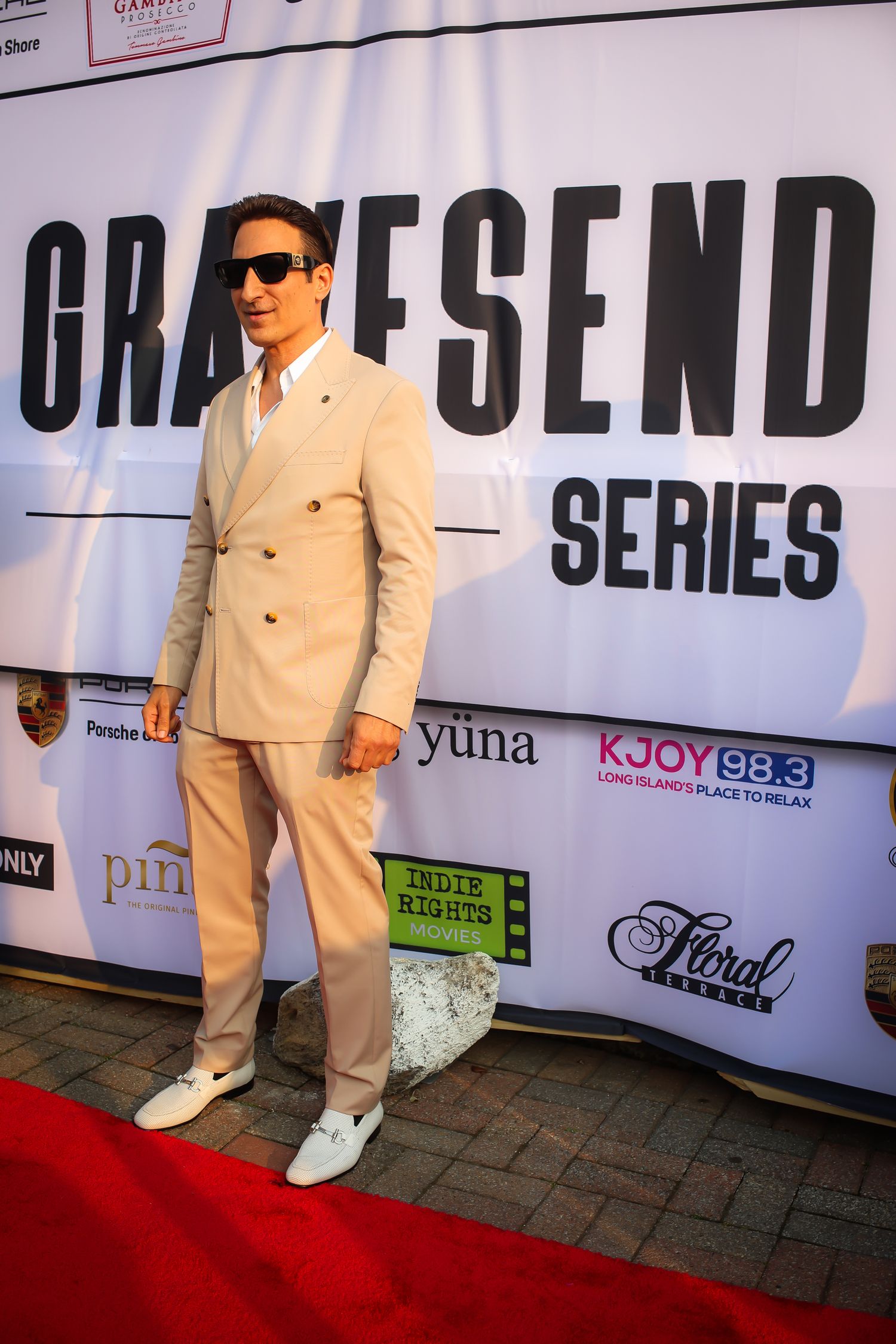 A person in a beige suit and sunglasses poses on a red carpet in front of a Graffsend Series backdrop.