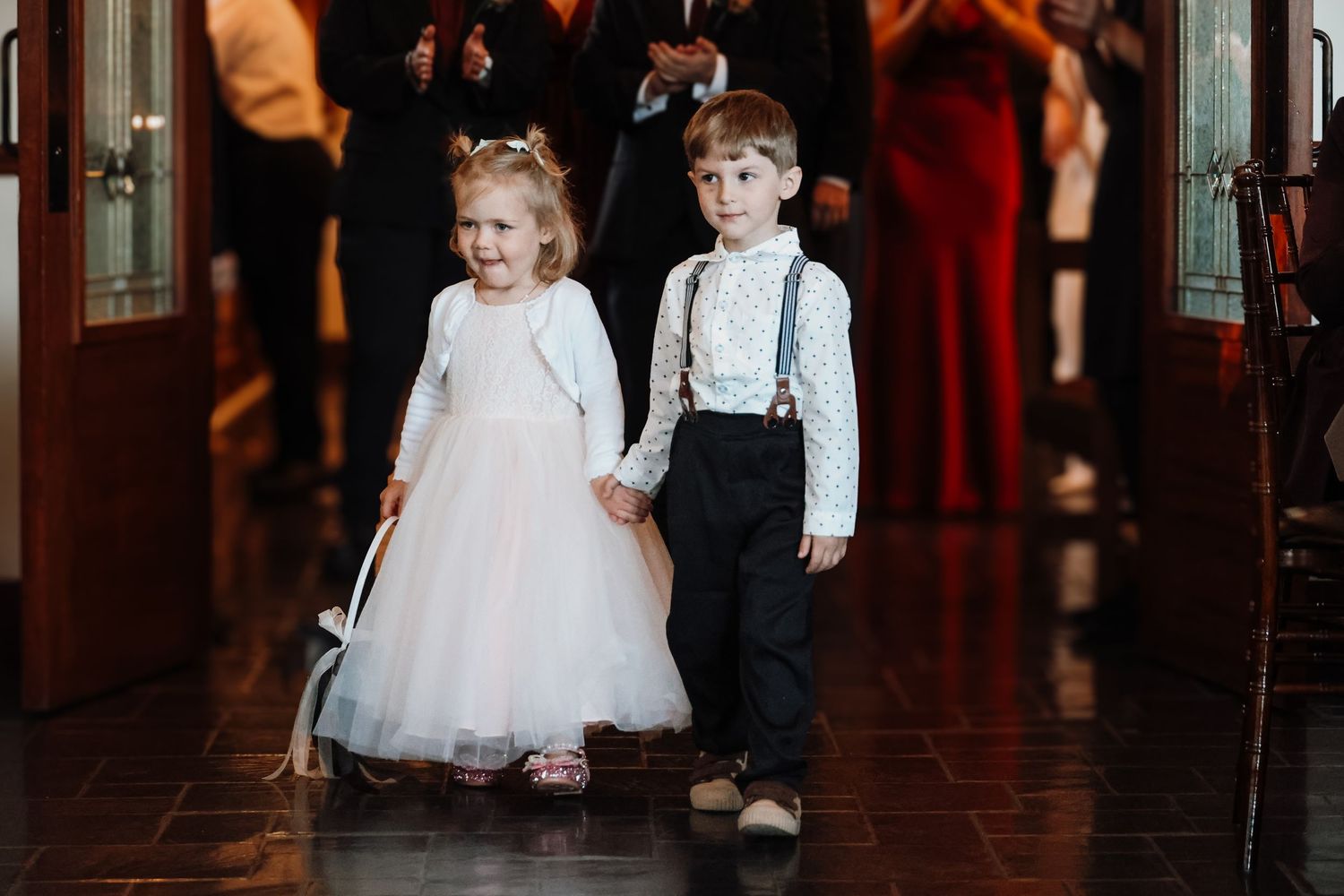 Two young children in formal attire walk down a dimly lit hallway during an elegant event.