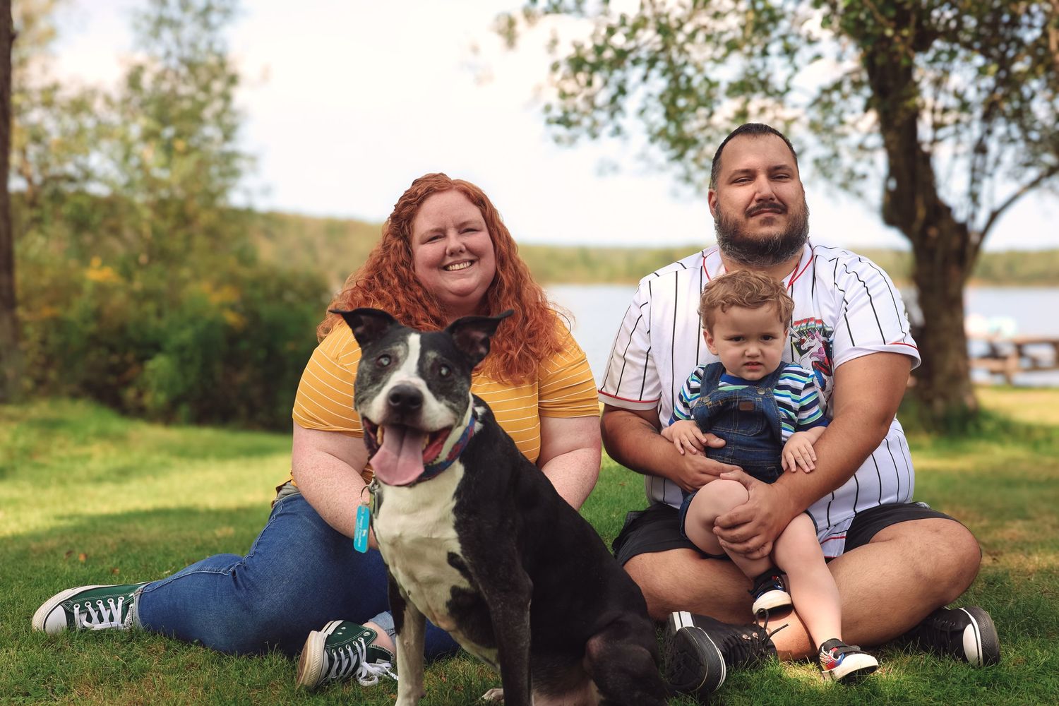 A family sits together with their black dog on a grassy hill during a sunny autumn day.