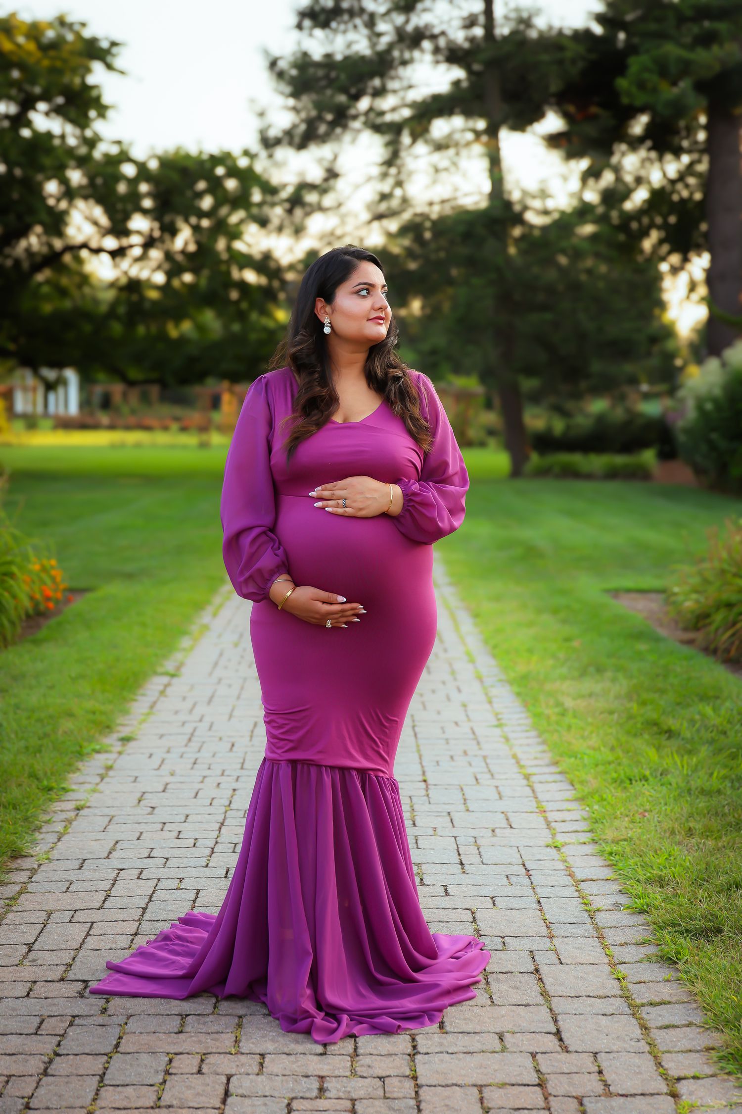 A figure in a flowing magenta dress poses gracefully on a stone path surrounded by lush green grass at sunset.