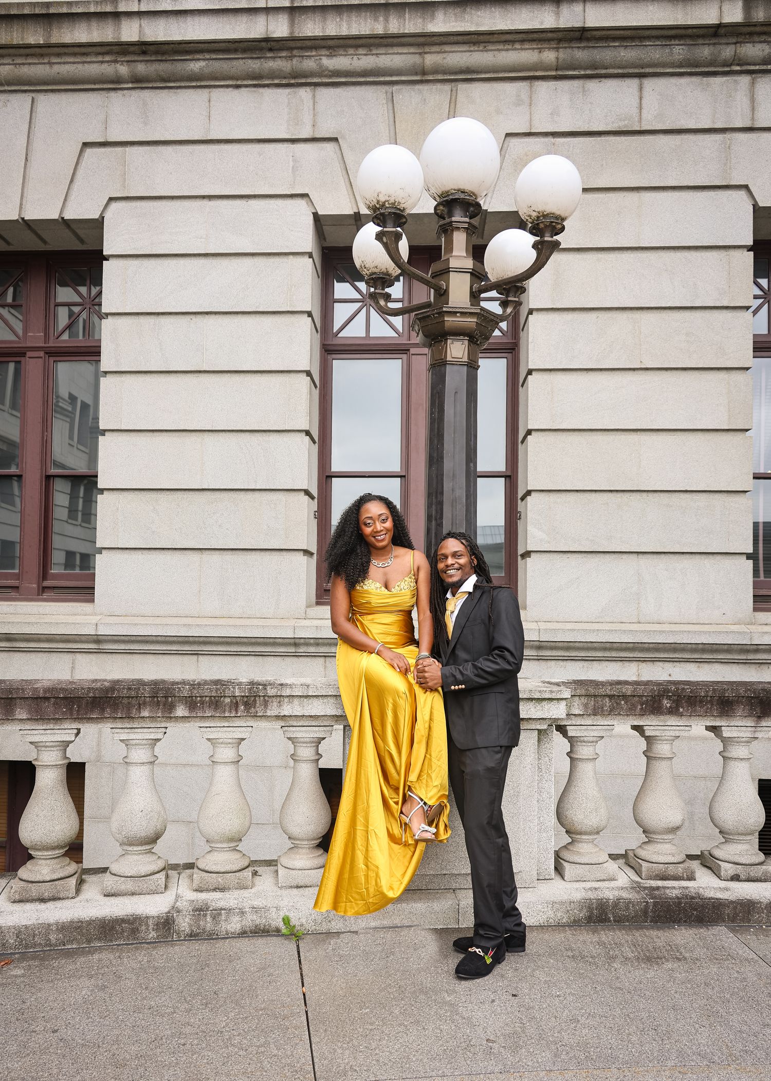 A couple in formal attire poses playfully on stone steps outside a historic building with white columns and globe lights.
