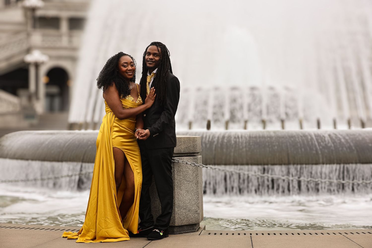A couple in a yellow dress and dark suit embrace lovingly near a fountain on a romantic evening.