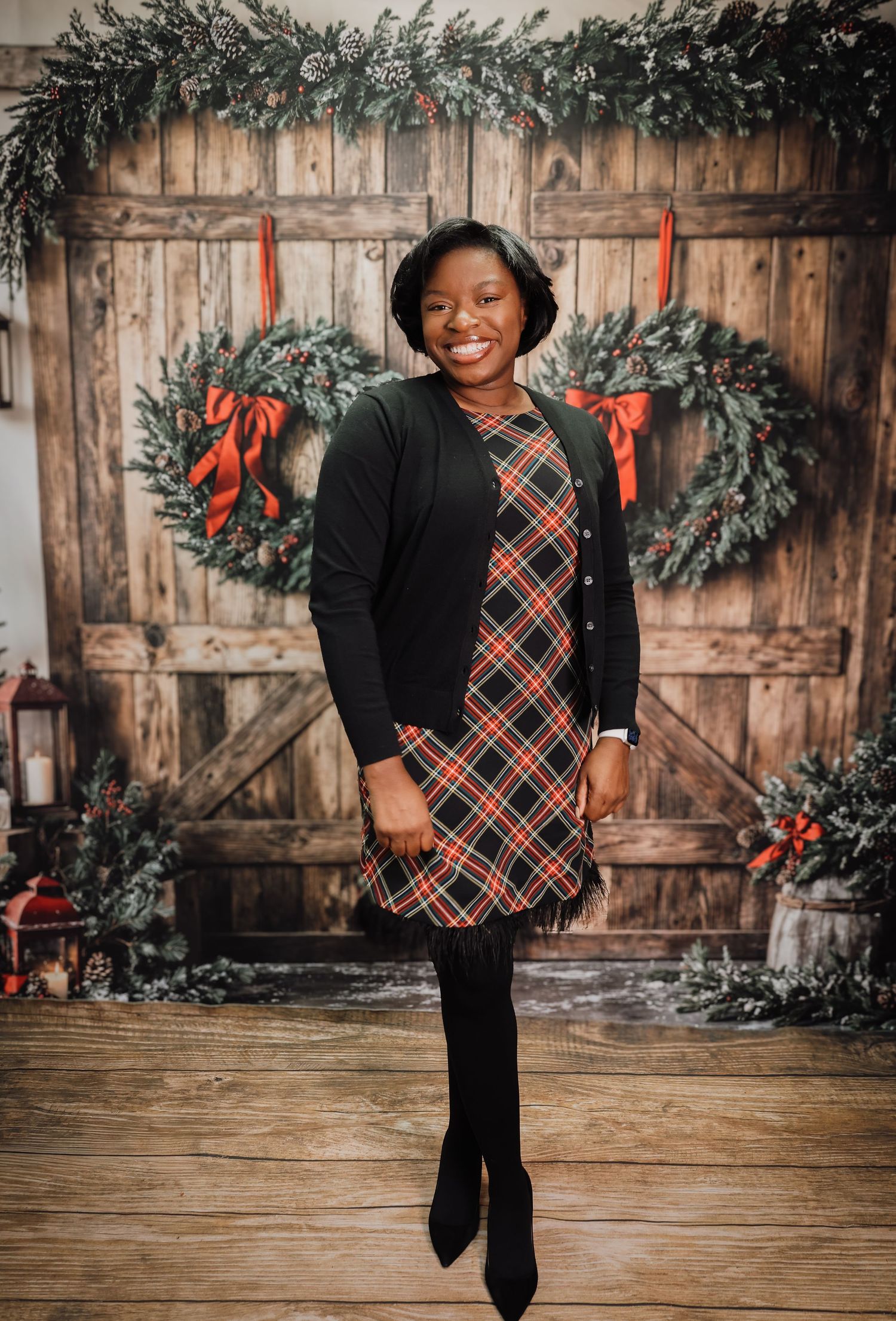 Woman in plaid dress and black cardigan smiling in front of rustic wooden doors with Christmas wreaths.