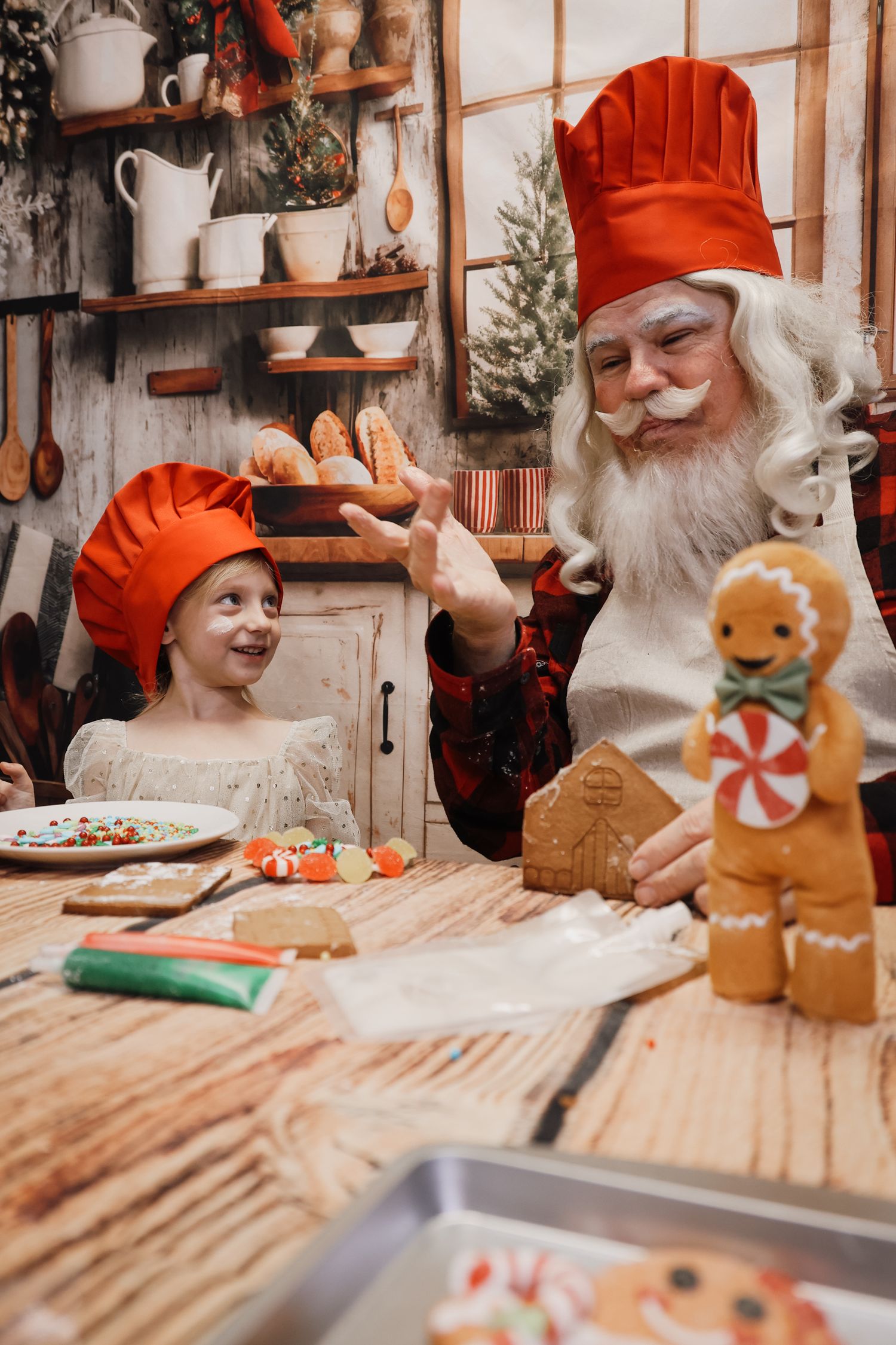 Santa Claus and a child in red chef hats decorate gingerbread cookies together in a festive Christmas kitchen.