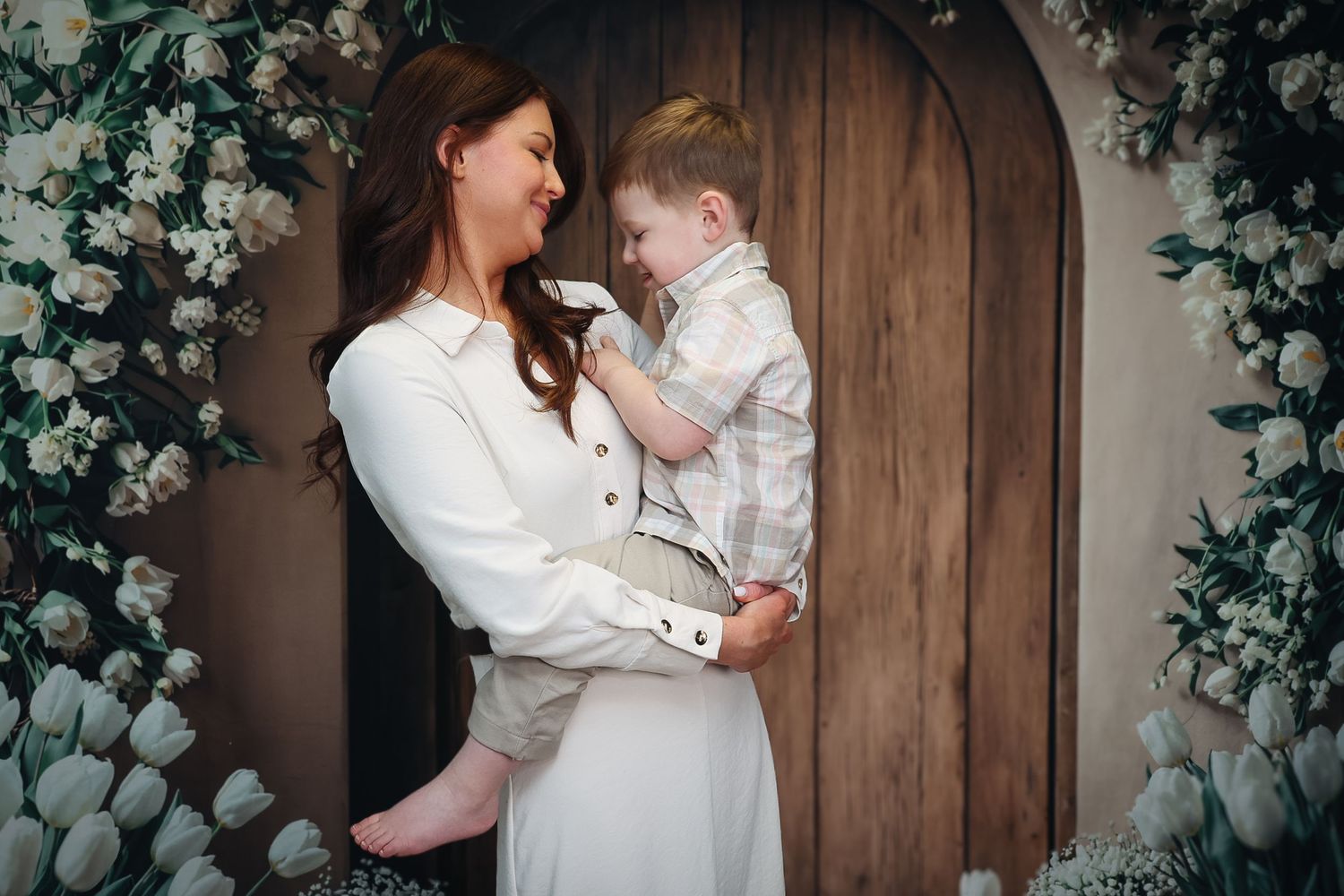 A mother in a white dress holds her young son in a suit near a floral arch with white tulips and roses.