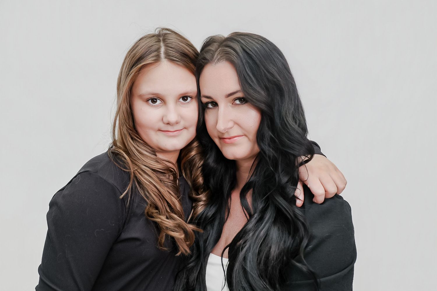 Two women with long hair in black jackets pose together smiling against a light gray background.