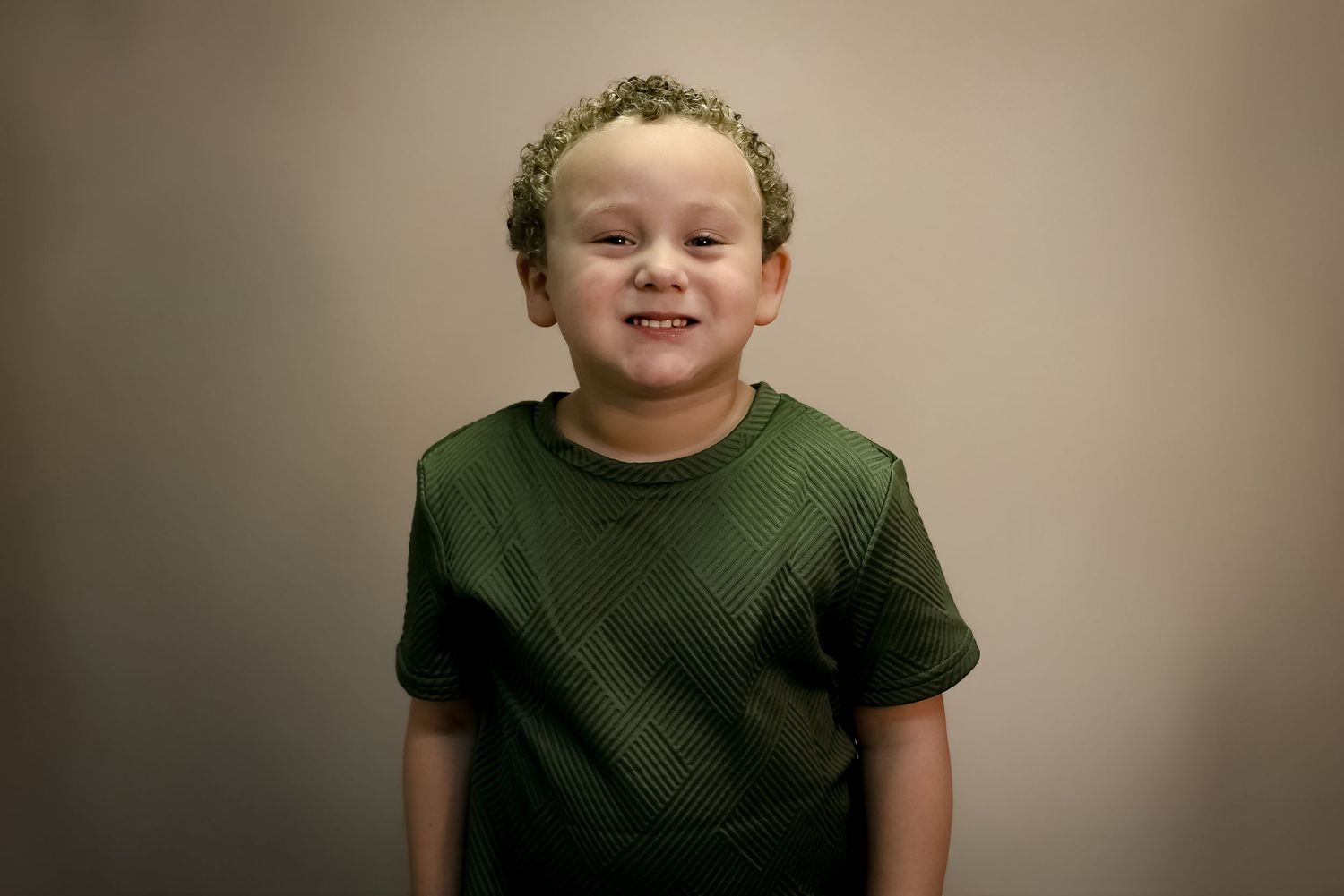 Smiling young boy with curly hair wearing a green t-shirt, posed against a neutral gray background.