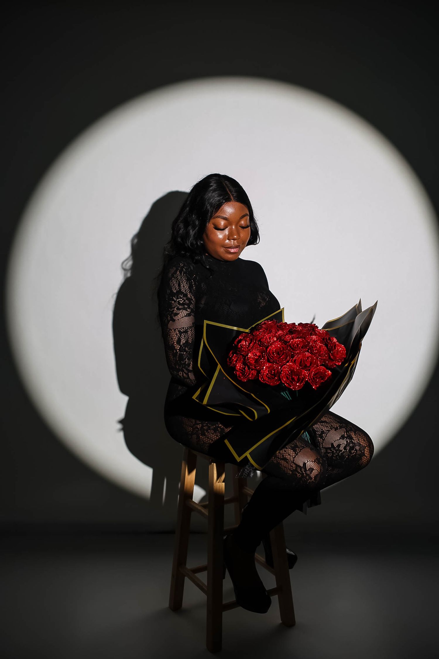 A Black woman in black lace sits on a stool holding a large red rose bouquet in black wrapping, lit by a circular spotlight.