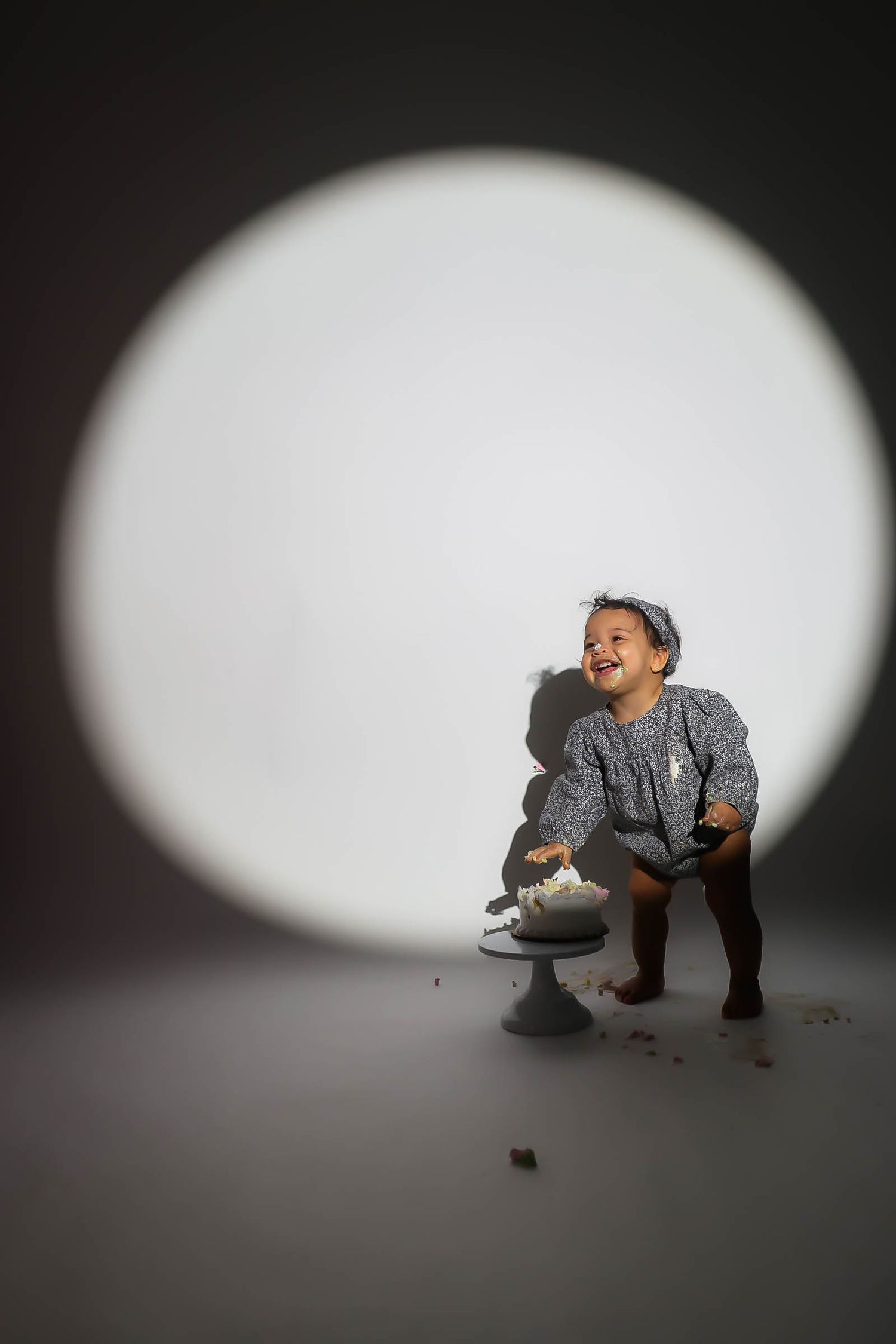 A laughing toddler in a gray dress smashes her first birthday cake under a bright circular spotlight.
