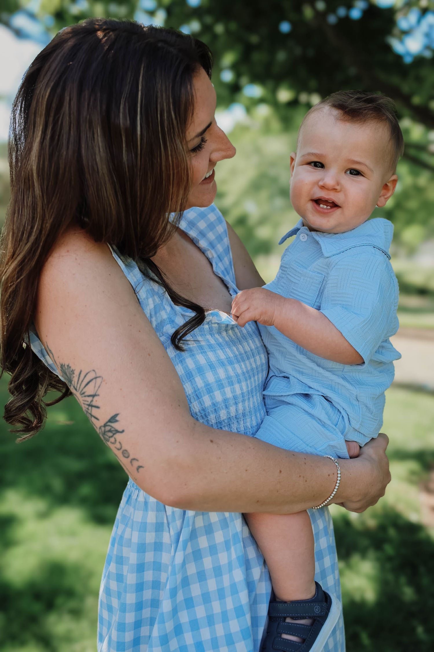 Mother in blue gingham dress holds smiling baby boy outdoors in green park.