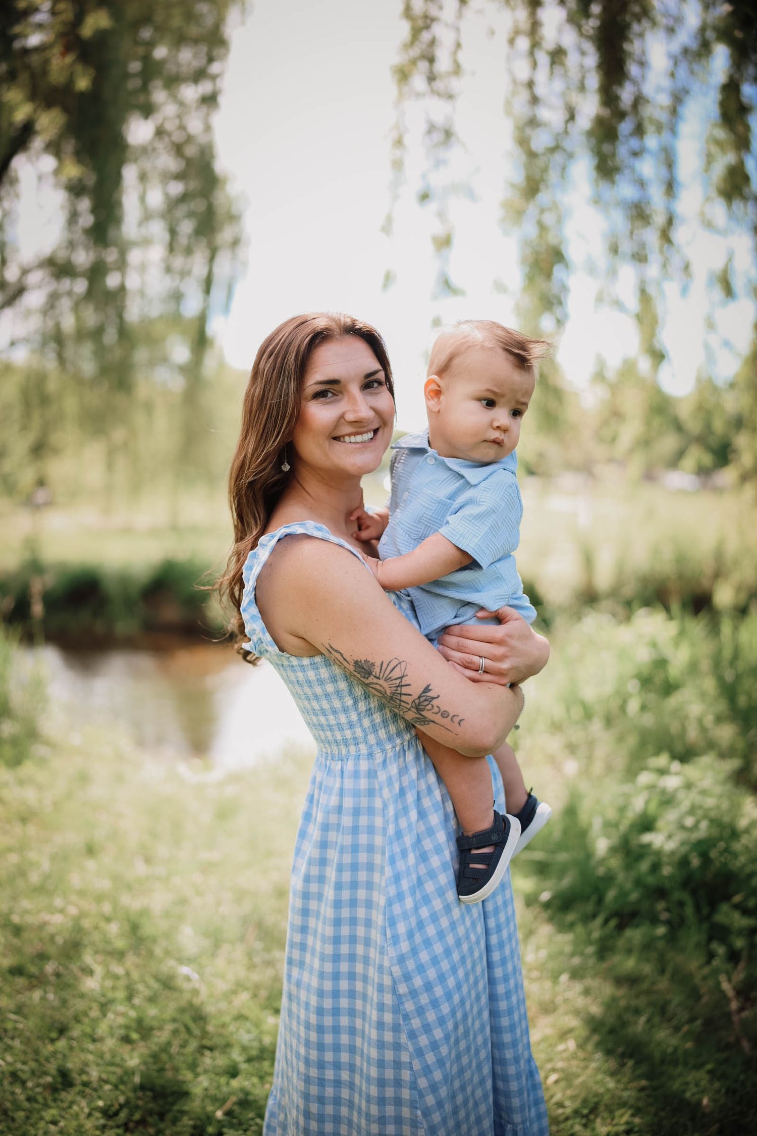 A smiling woman in a blue gingham dress holds a baby boy outdoors near a pond surrounded by lush green trees.
