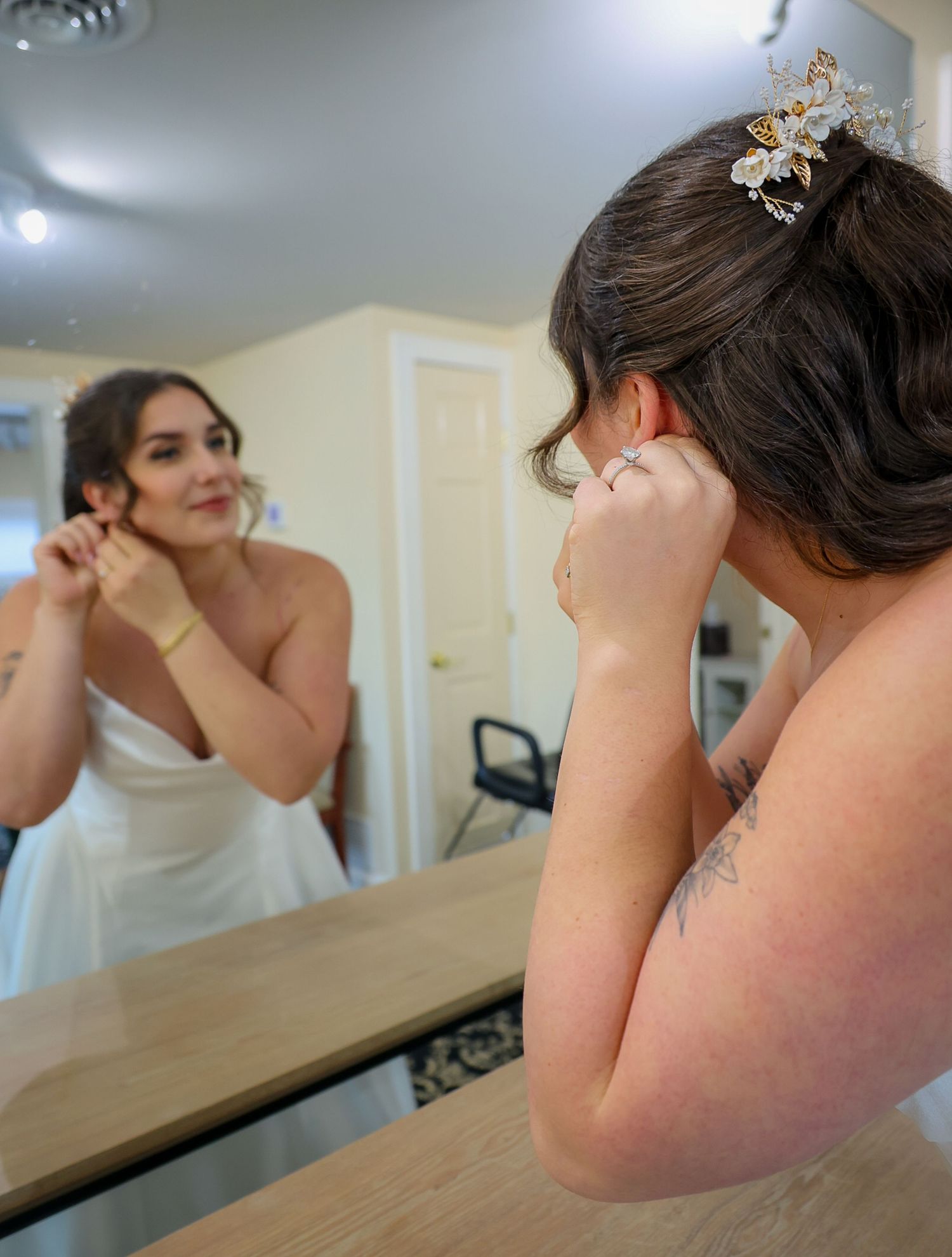 Bride in white gown adjusting earring before mirror, wearing gold butterfly hair comb and showing shoulder tattoo.