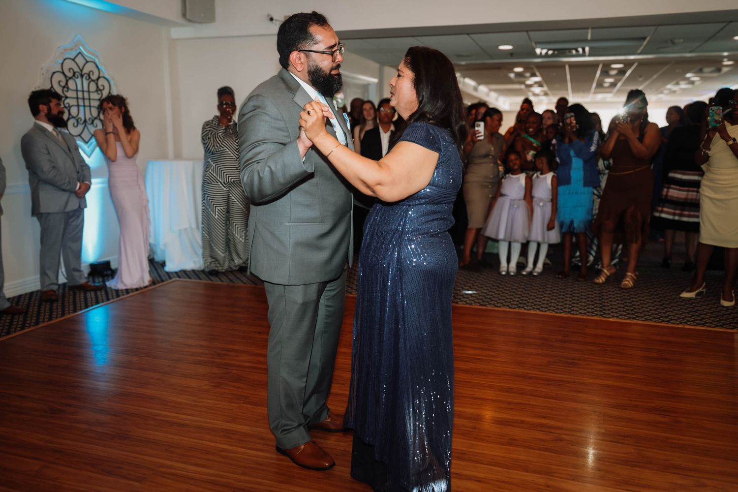 A couple shares a mother-son dance at a wedding reception in a elegant ballroom with guests watching in the background.