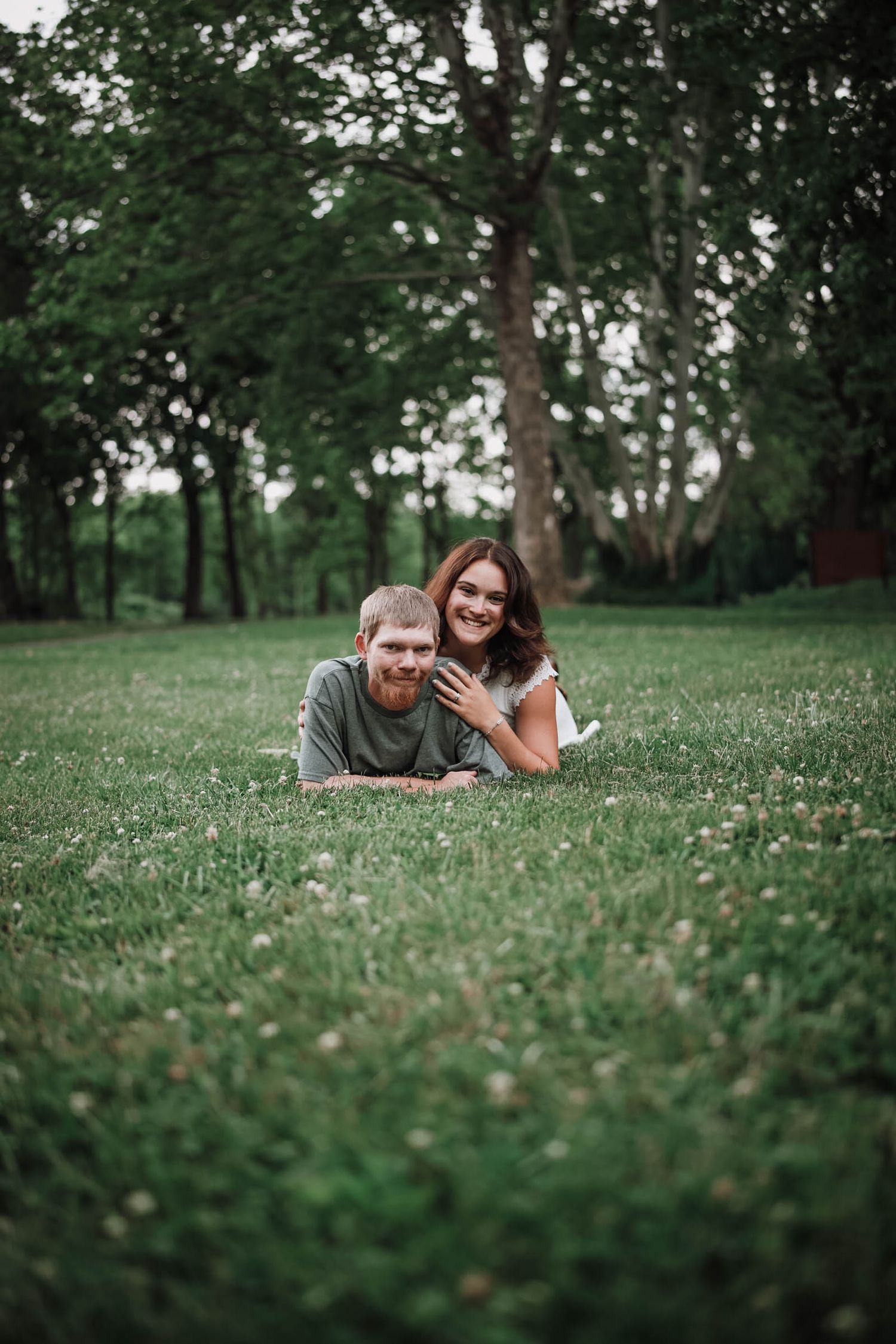 Happy couple lying on lush green grass in a park, smiling together surrounded by trees.