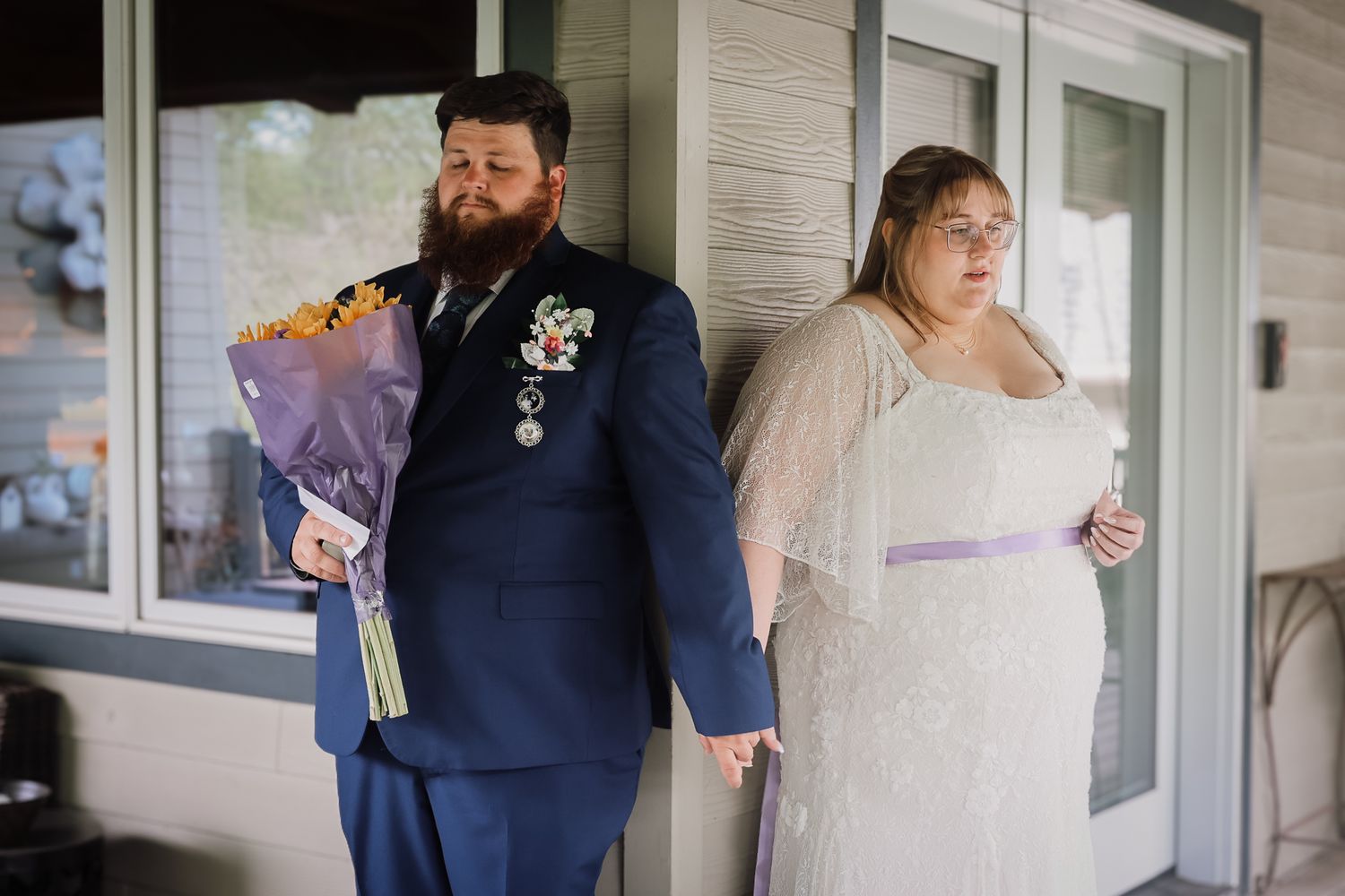 Groom in navy suit holding sunflower bouquet stands with bride in white gown on porch before wedding ceremony.