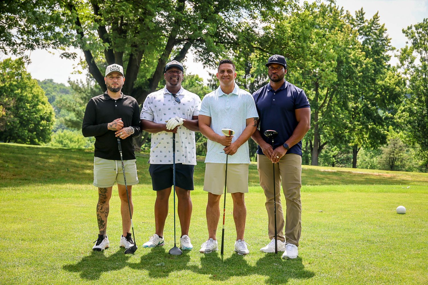 Four men posing together on a golf course, holding clubs, surrounded by lush green trees on a sunny day.