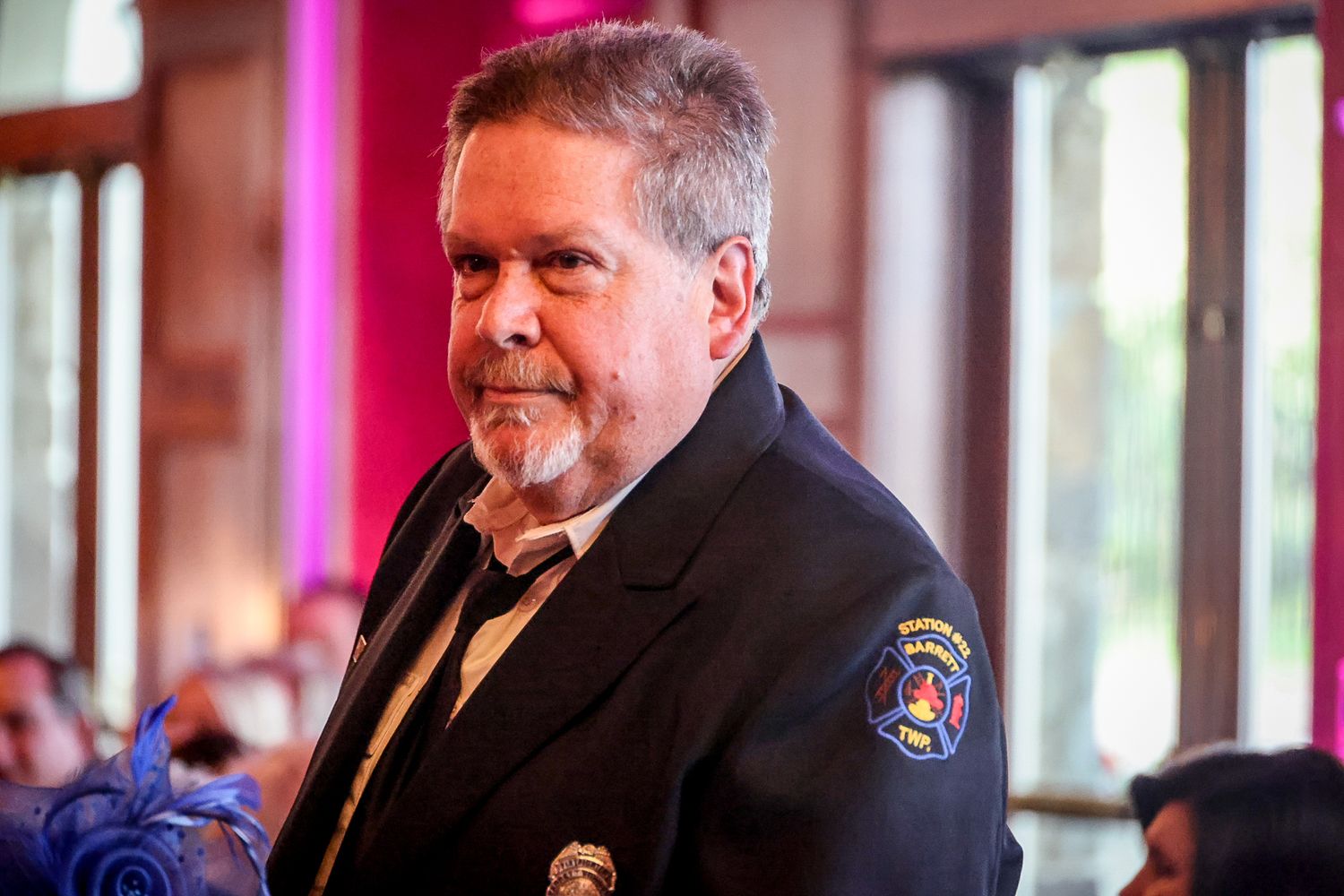 Middle-aged man in dark uniform jacket with badge at formal indoor event with pink ambient lighting.