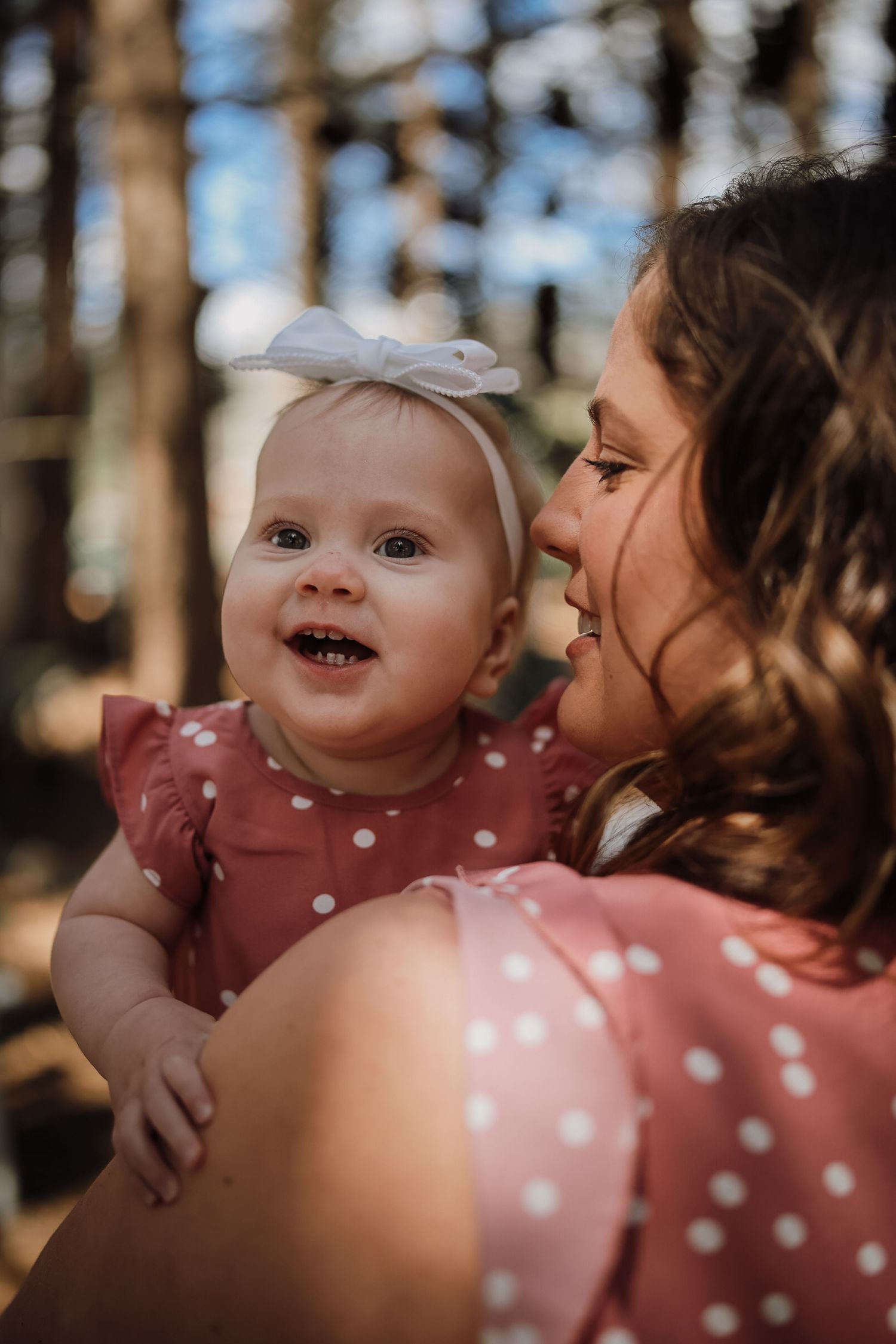A smiling baby in a pink polka dot dress with a white bow headband being held by a woman outdoors.