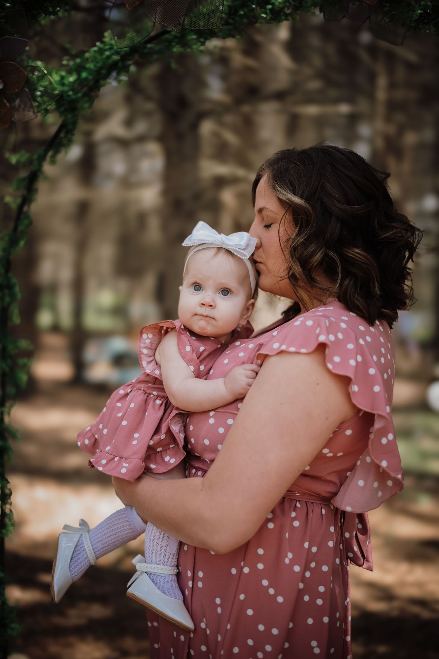 Mother in pink polka dot dress kissing baby girl in matching outfit with white bow headband in forest.