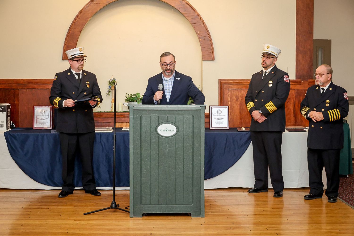 Four officials in uniforms stand at a podium during a formal fire department awards ceremony indoors.