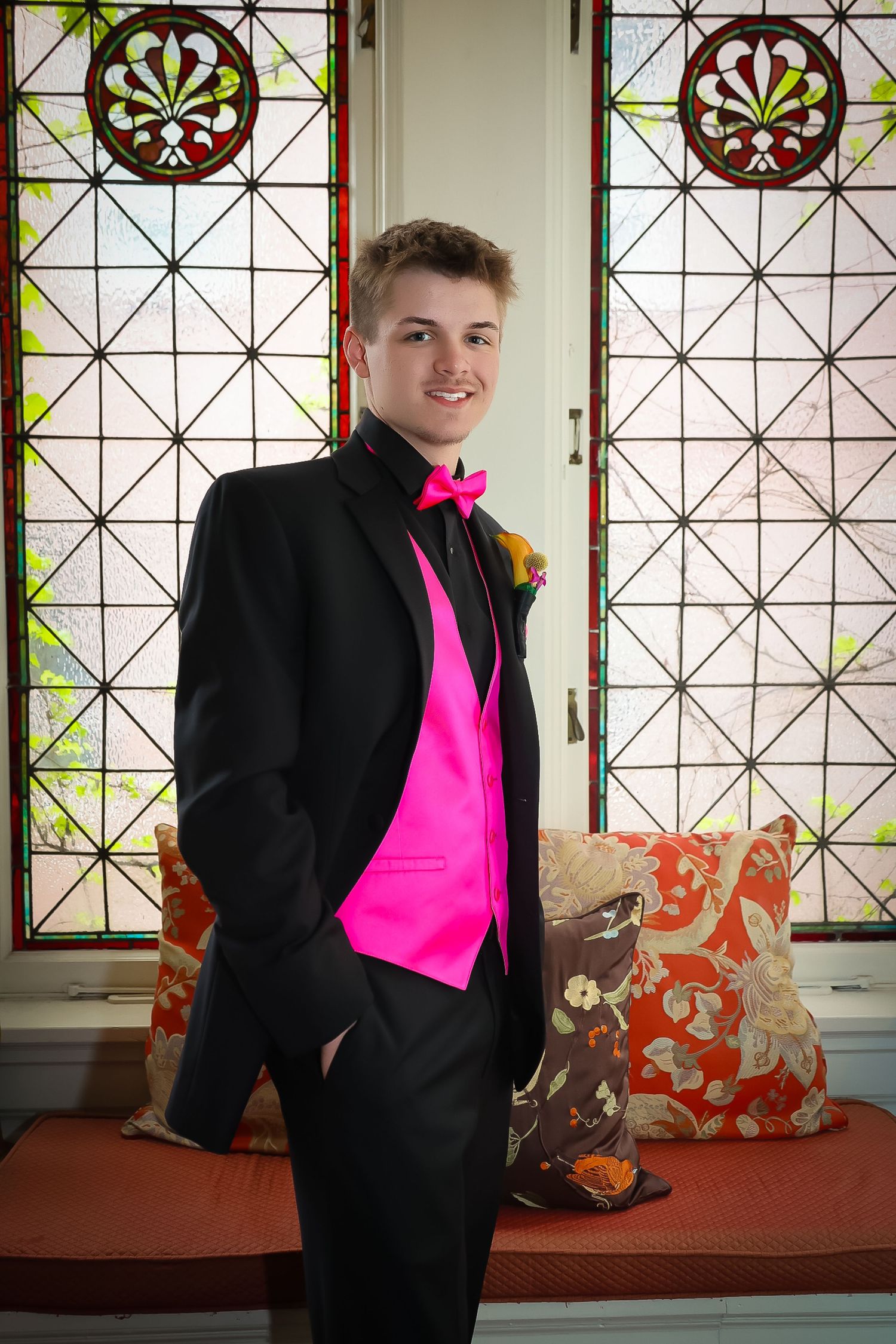 Young man in black tuxedo with hot pink vest and bow tie poses before stained glass windows at prom.