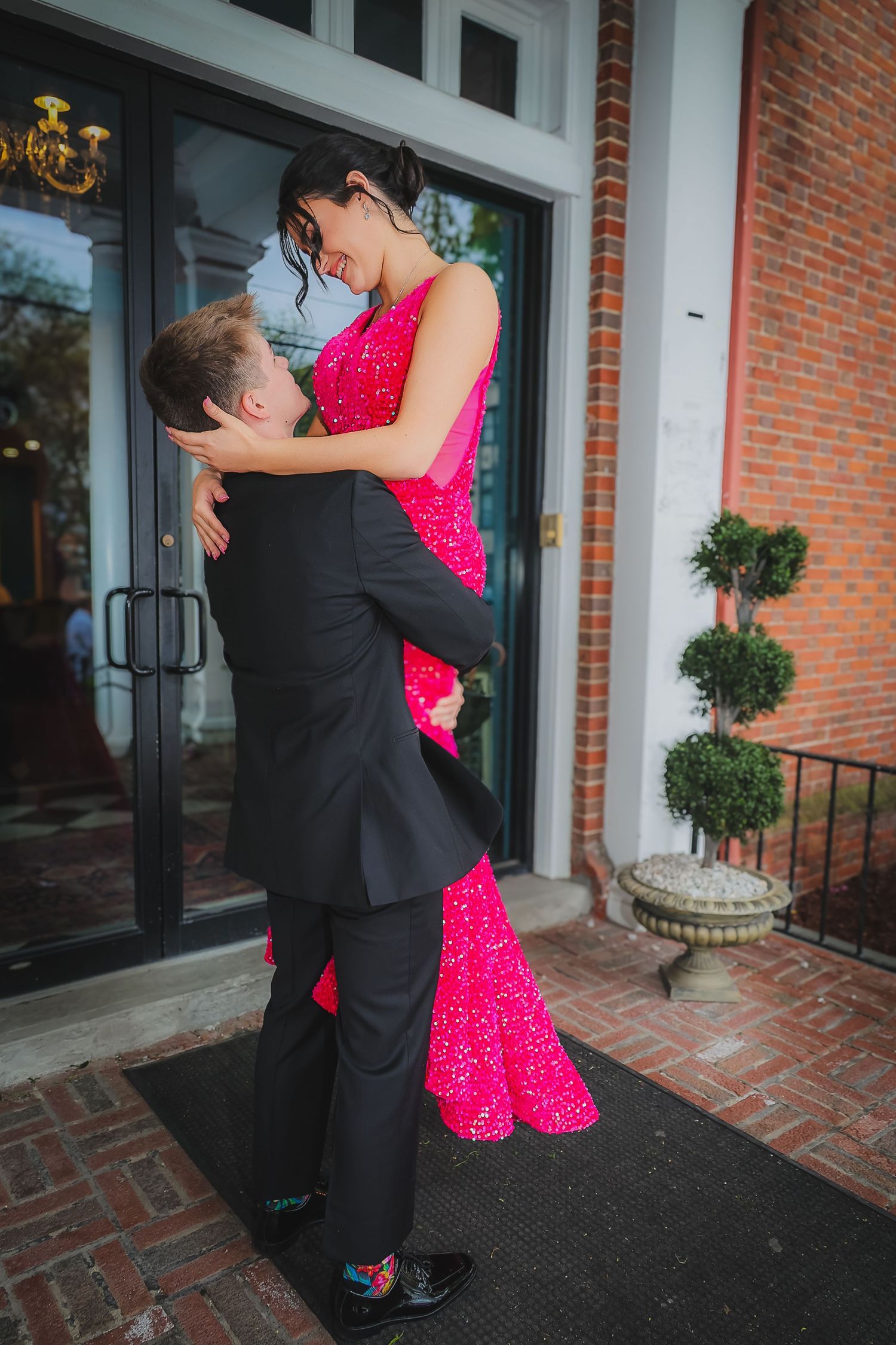 Couple embracing at prom, woman in pink sequin gown lifted by man in black tuxedo outside brick building.