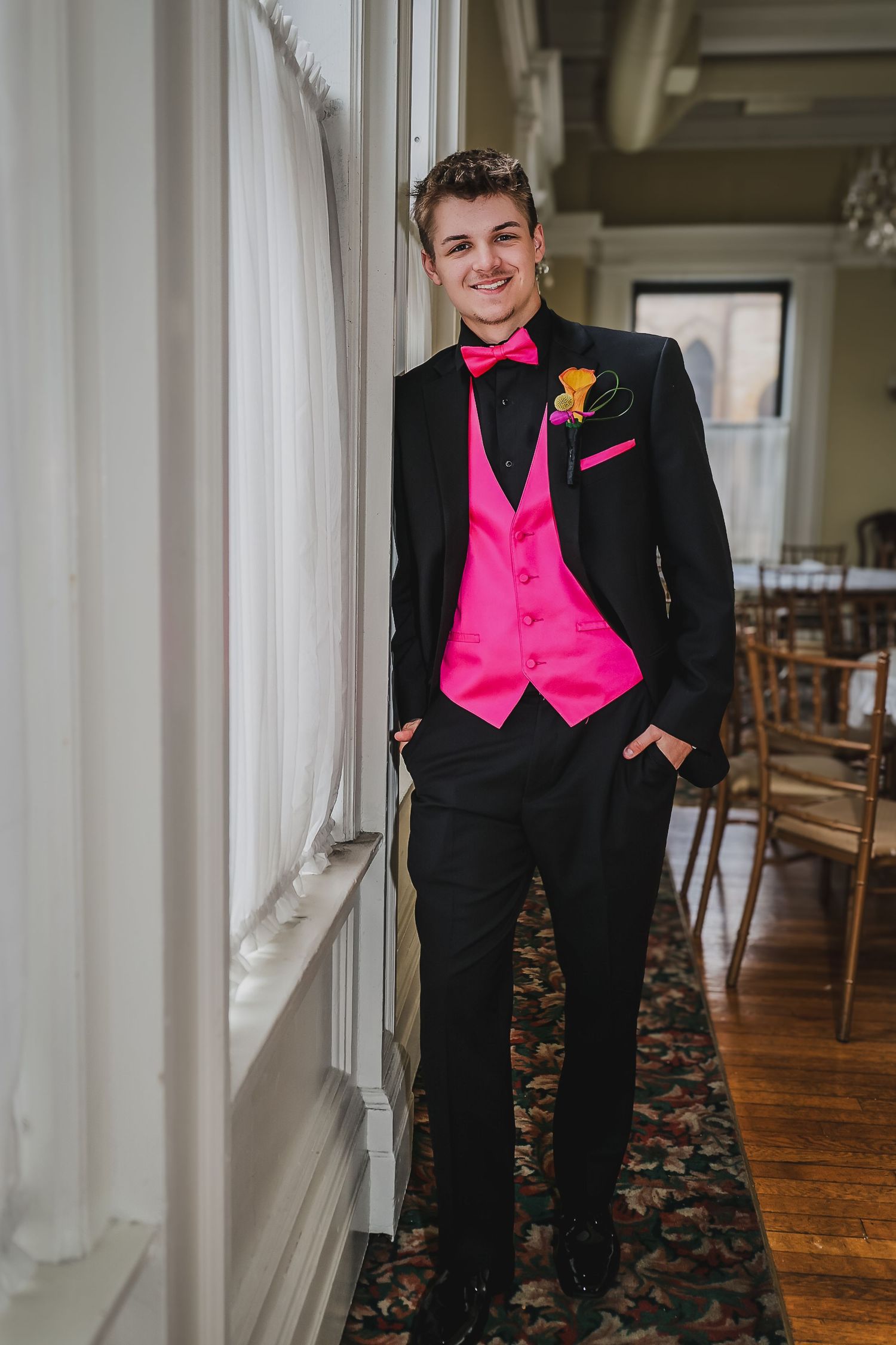 Young man in black tuxedo with hot pink vest, bow tie, and boutonniere posing by window at formal event.