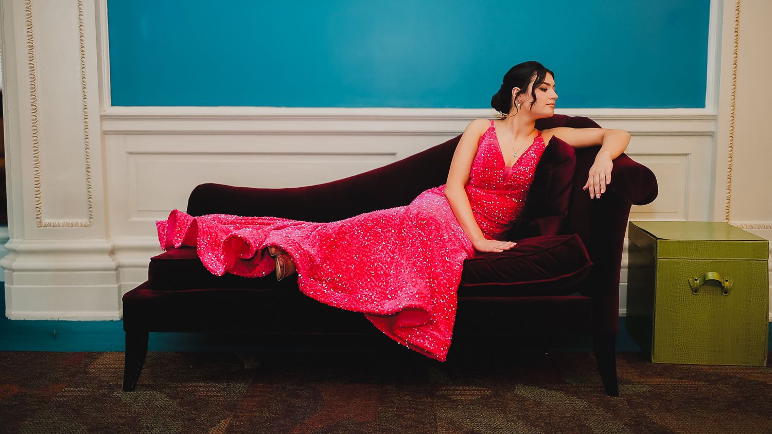 Woman in a pink sequin gown reclining on a dark velvet chaise lounge against a teal wall.