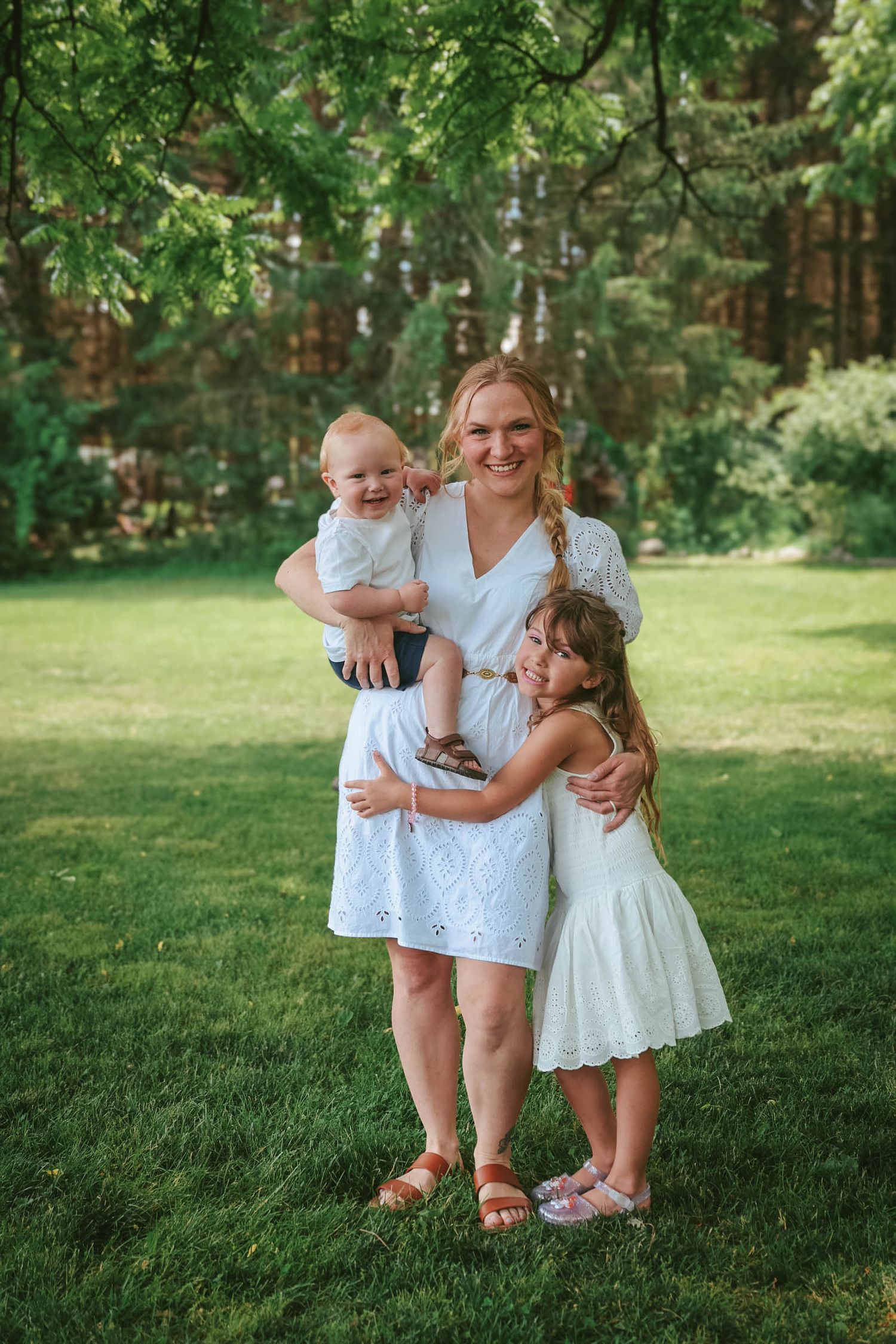A smiling woman in a white dress holds a baby while a young girl in white hugs her outdoors on green grass.