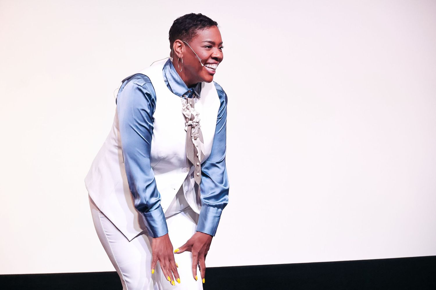 Young man in blue satin shirt and white vest laughing on stage under bright lights.