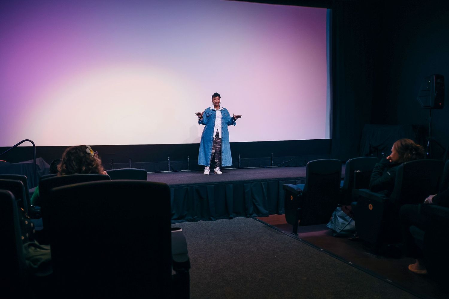 Person presenting on stage in a darkened cinema with a glowing pink and white screen backdrop.