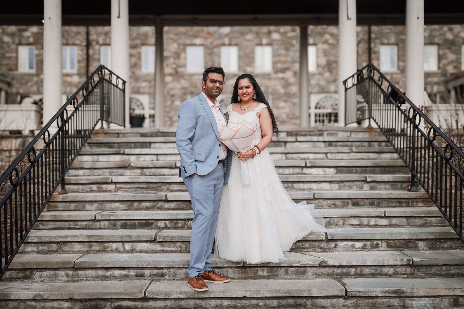 A couple in wedding attire stands on grand stone steps with iron railings outside a historic building.