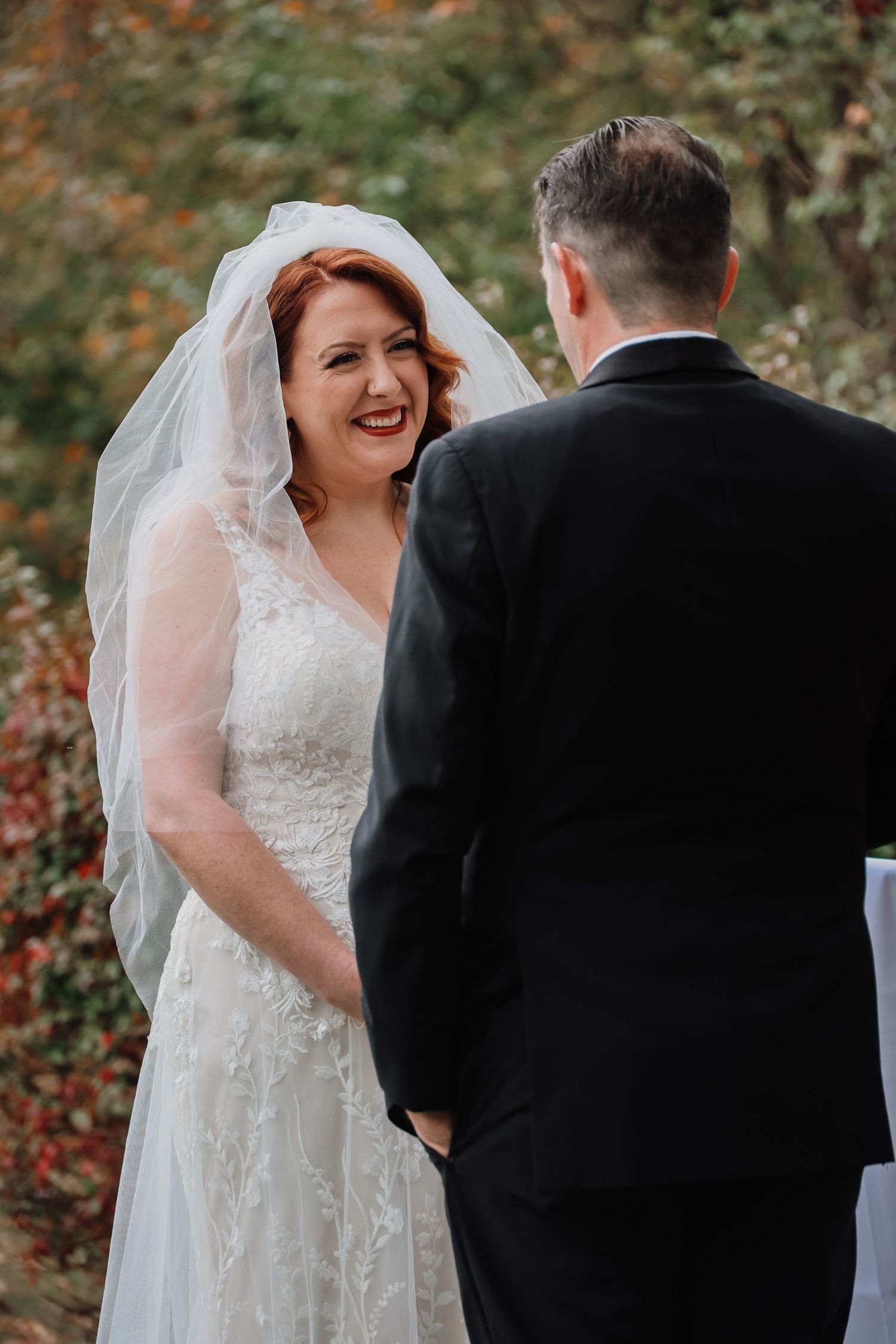 A bride in a white lace gown and veil smiles joyfully during an outdoor autumn wedding ceremony.