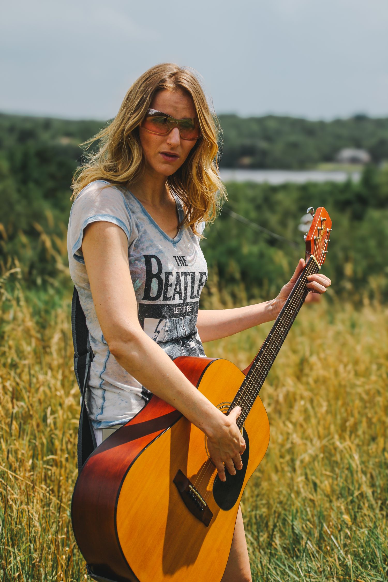 Musician in gray tank top playing acoustic guitar in golden summer field with green hills in background.