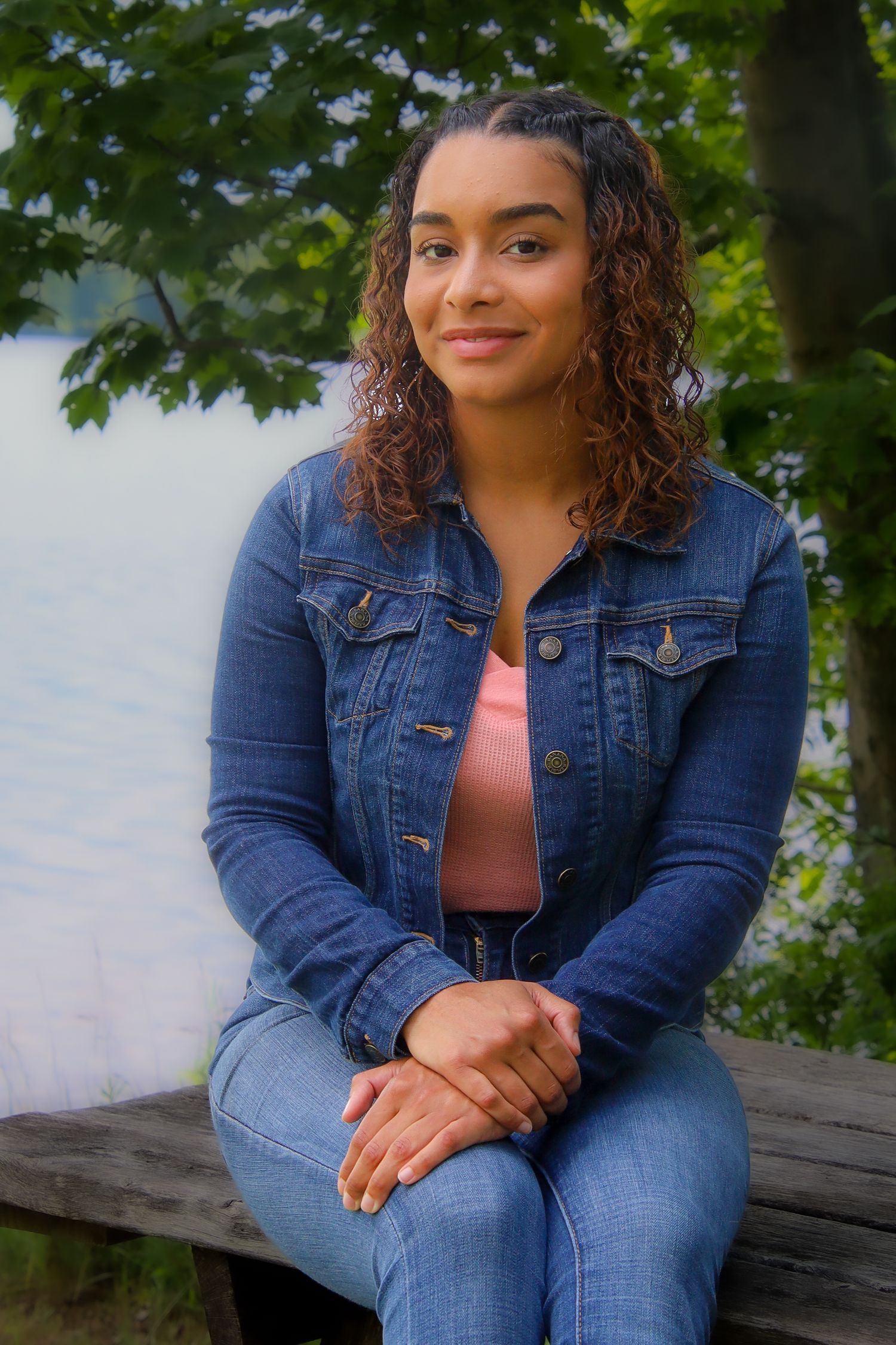 Young woman in denim jacket sits outdoors by water on sunny day with green foliage in background.