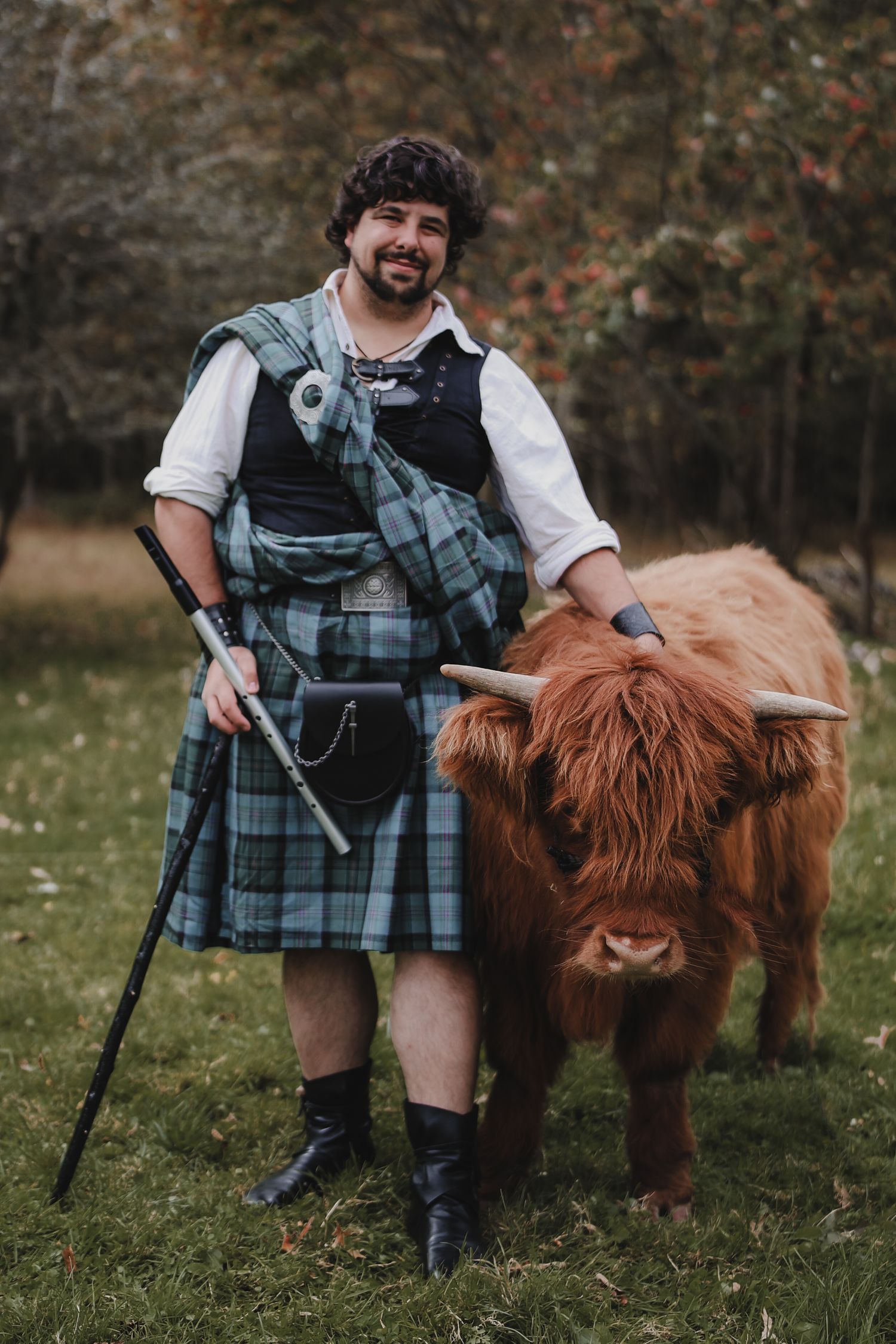 A person in traditional Scottish attire poses with a Highland cow in a grassy field during autumn.