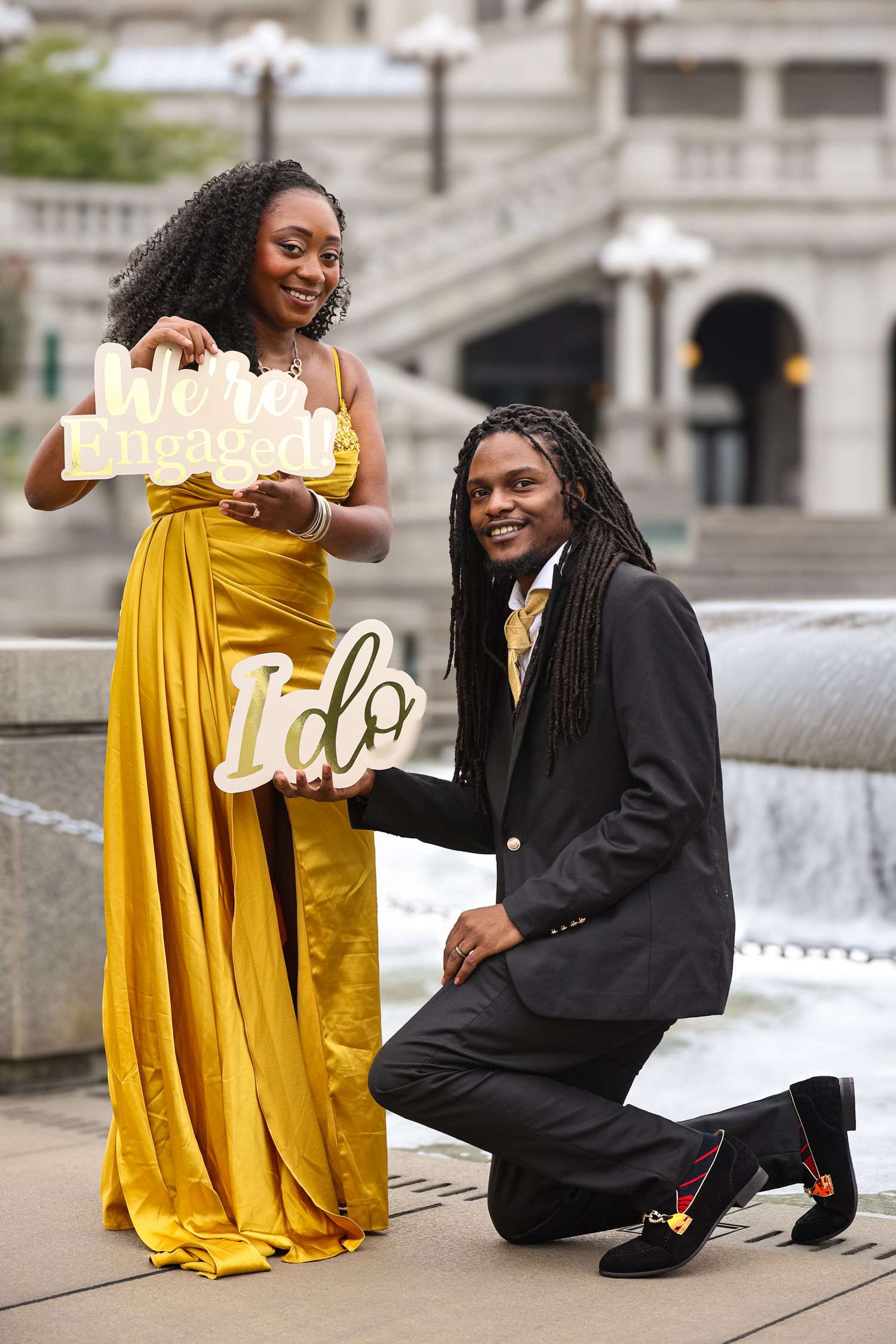 Two people pose together in a photo session, one wearing a yellow dress and one in black attire near stone steps.
