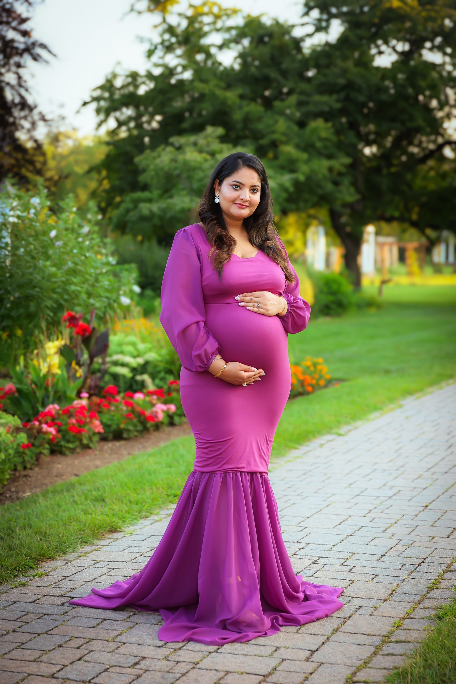 A pregnant figure in a flowing purple maternity gown poses in a garden with red flowers and a stone path.