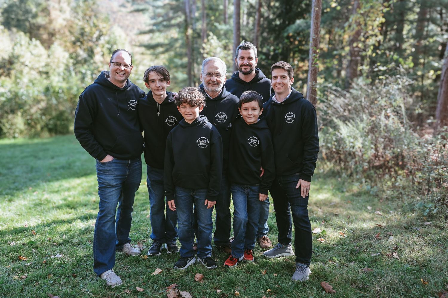 Group of people wearing dark jackets pose together outdoors in a wooded natural setting.