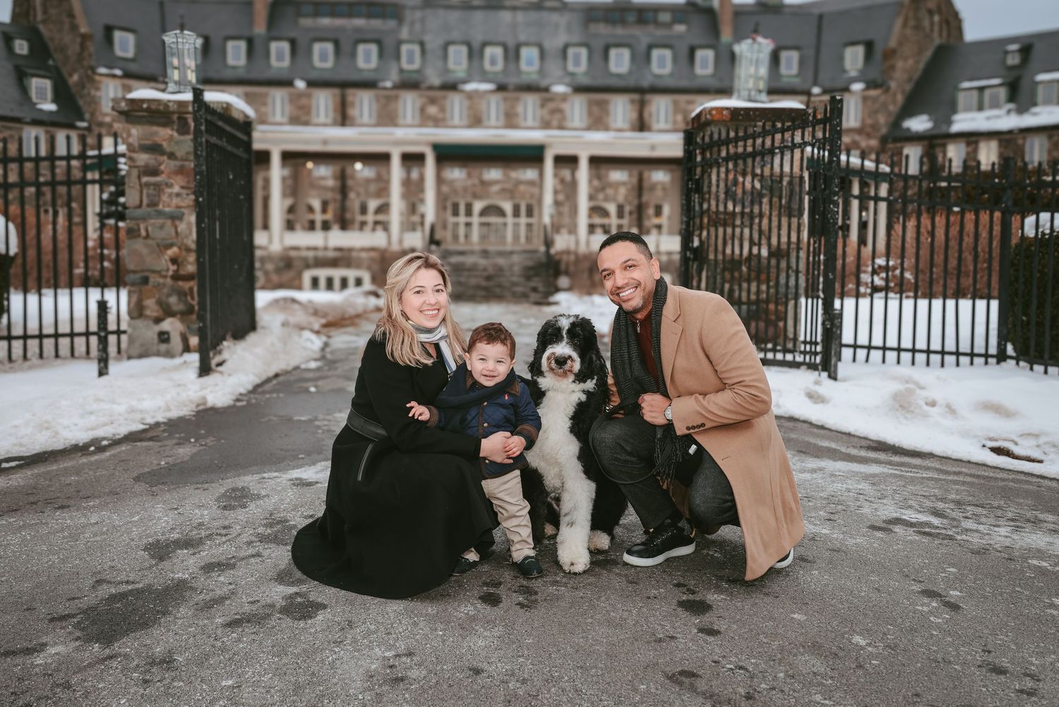 A family poses together on a snowy winter day in front of an elegant mansion with ornate iron gates.