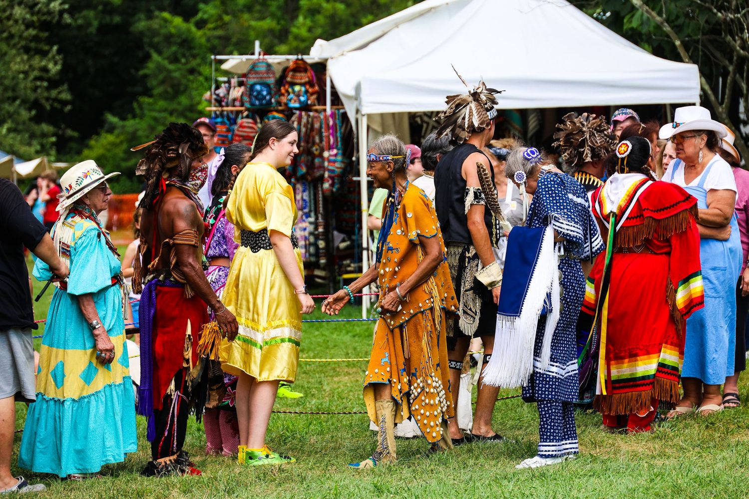 A line of people in colorful traditional Native American regalia gather outdoors for a ceremonial dance event.