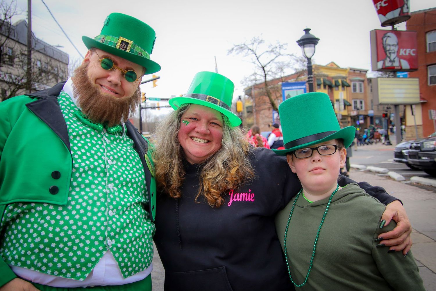 Three people wearing green St. Patrick's Day hats and festive attire smile together at a street celebration.