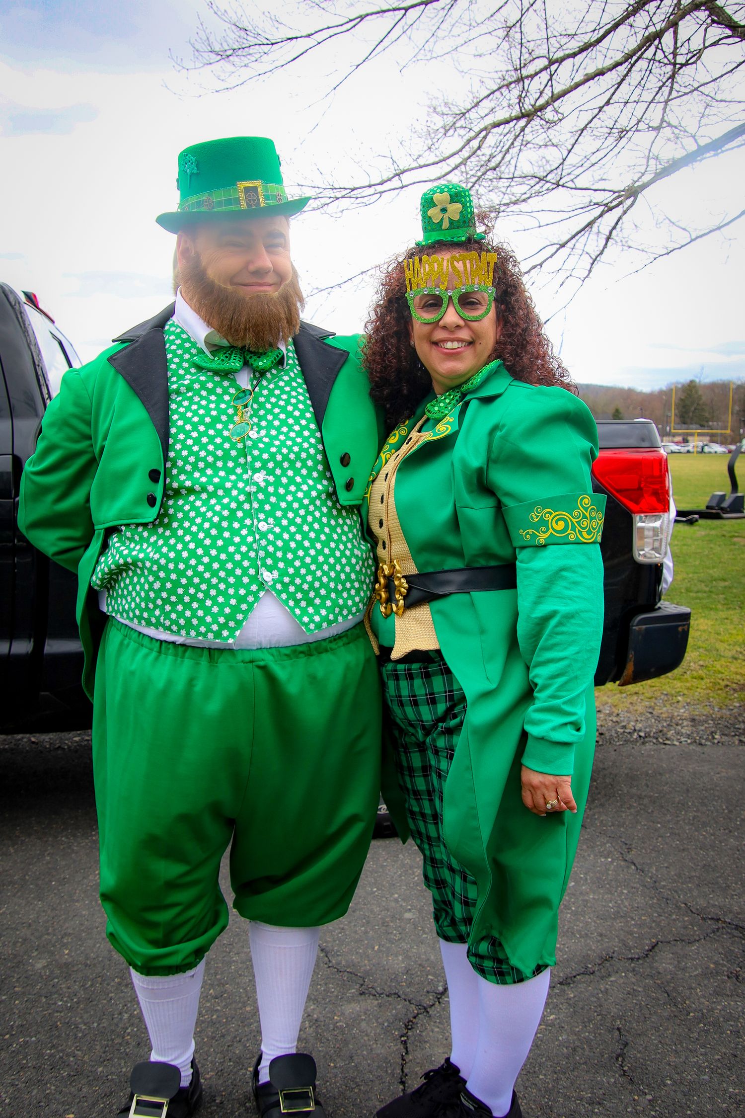 Two people dressed in festive green St. Patrick's Day costumes with shamrock accessories pose together outdoors.