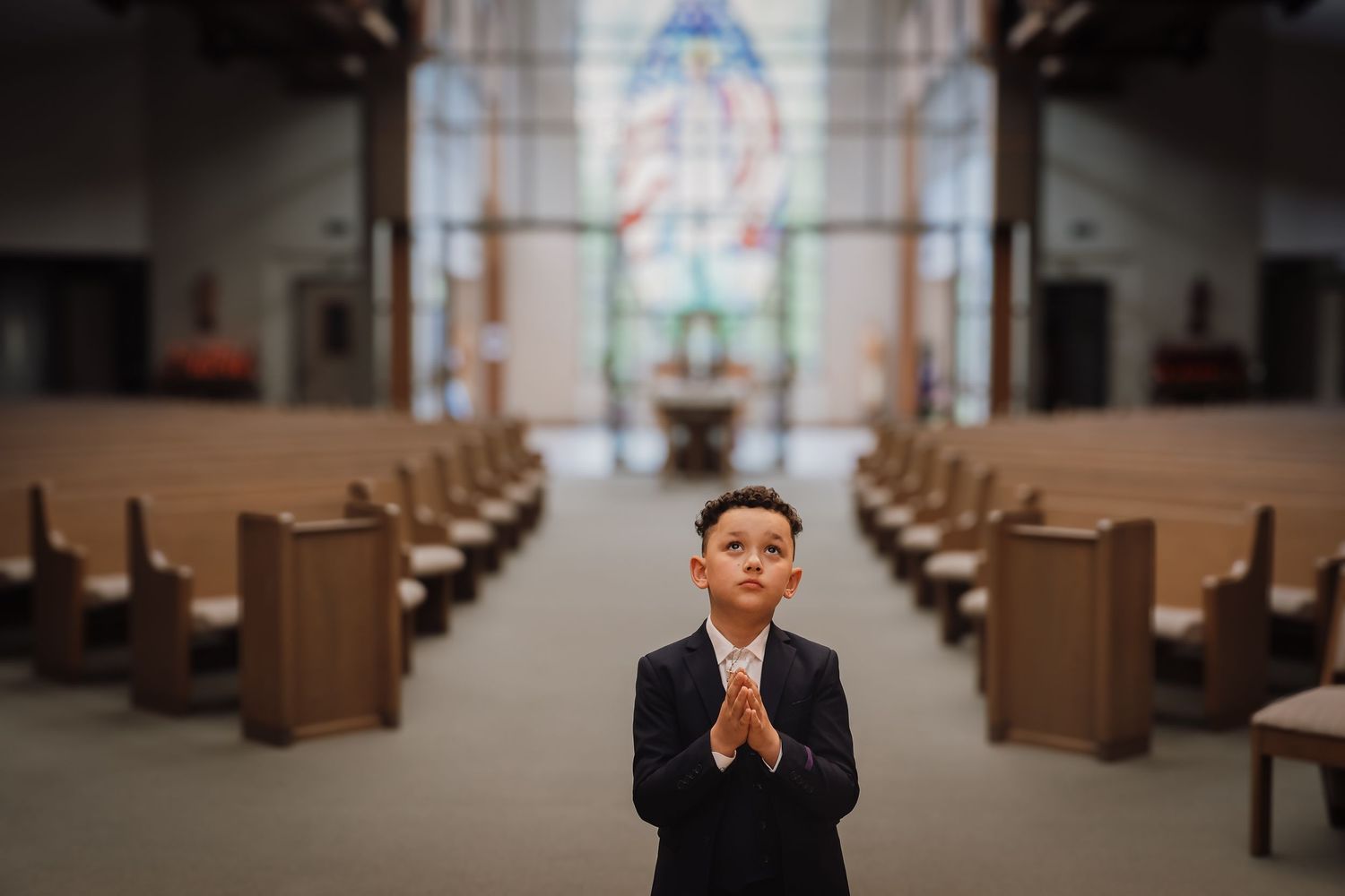 A person in a black suit stands in prayer at the center aisle of an empty church with stained glass windows.