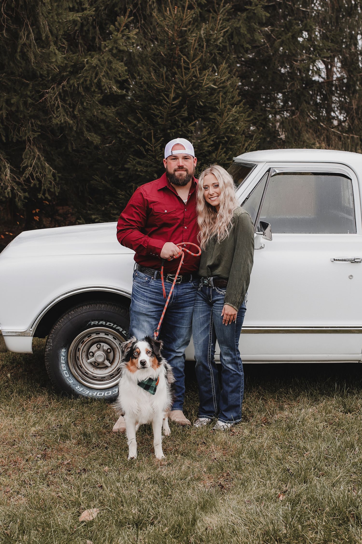 A couple poses with their Australian Shepherd dog next to a vintage white car in a grassy outdoor setting.