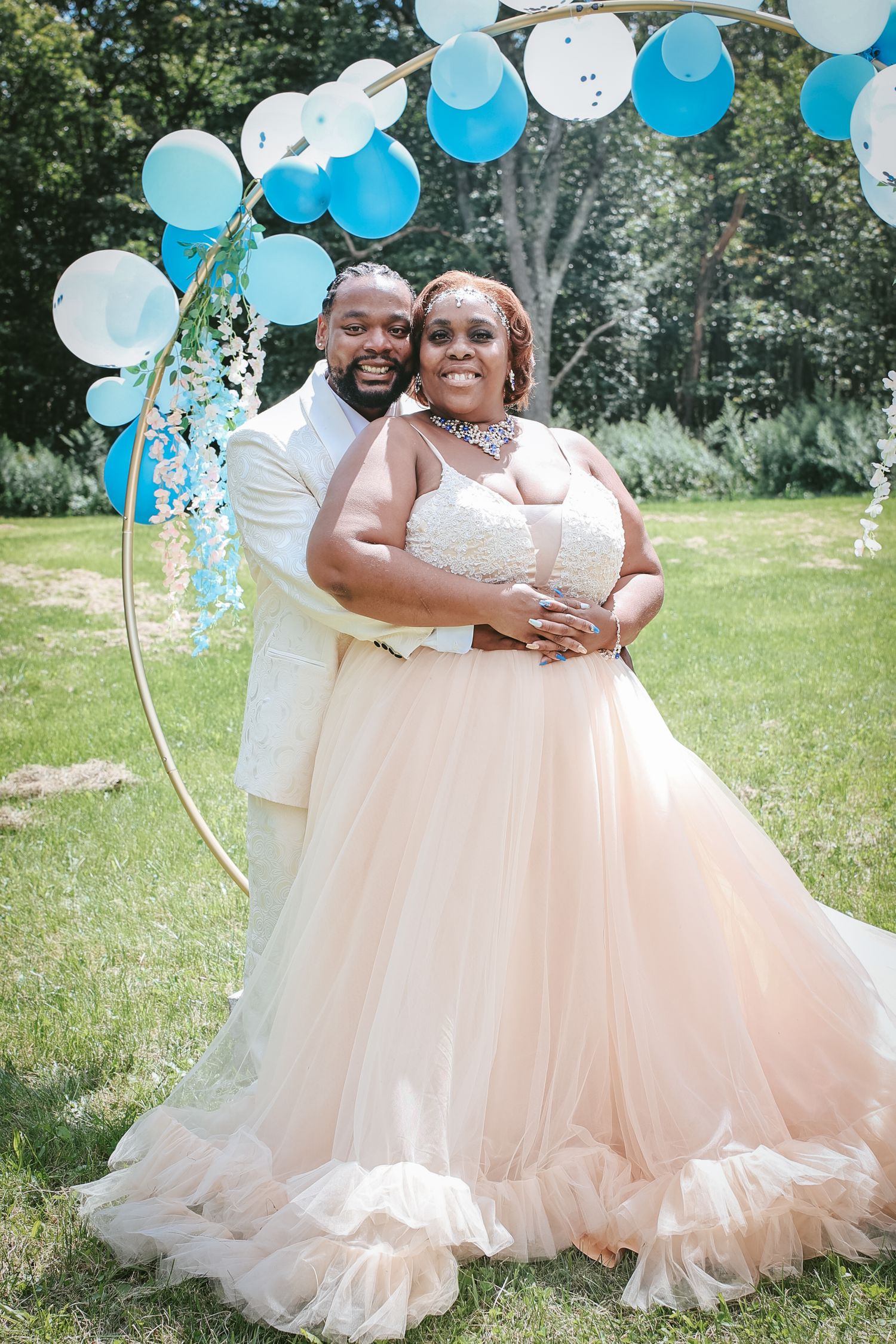 Couple in matching blush pink tulle gowns poses outdoors with blue and white balloons during celebration photoshoot.