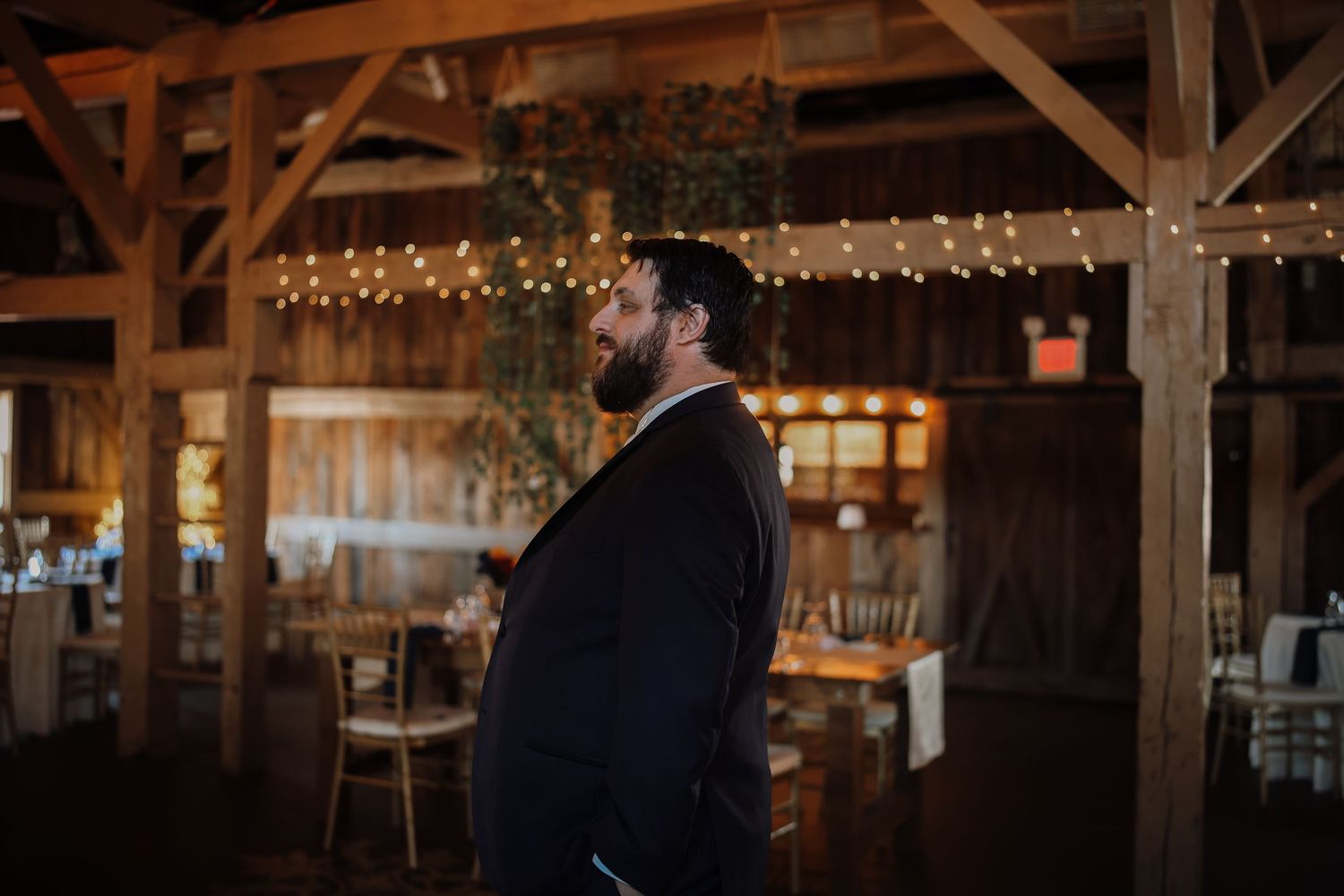 A groom in a black suit stands in a rustic barn venue decorated with string lights for a wedding reception.