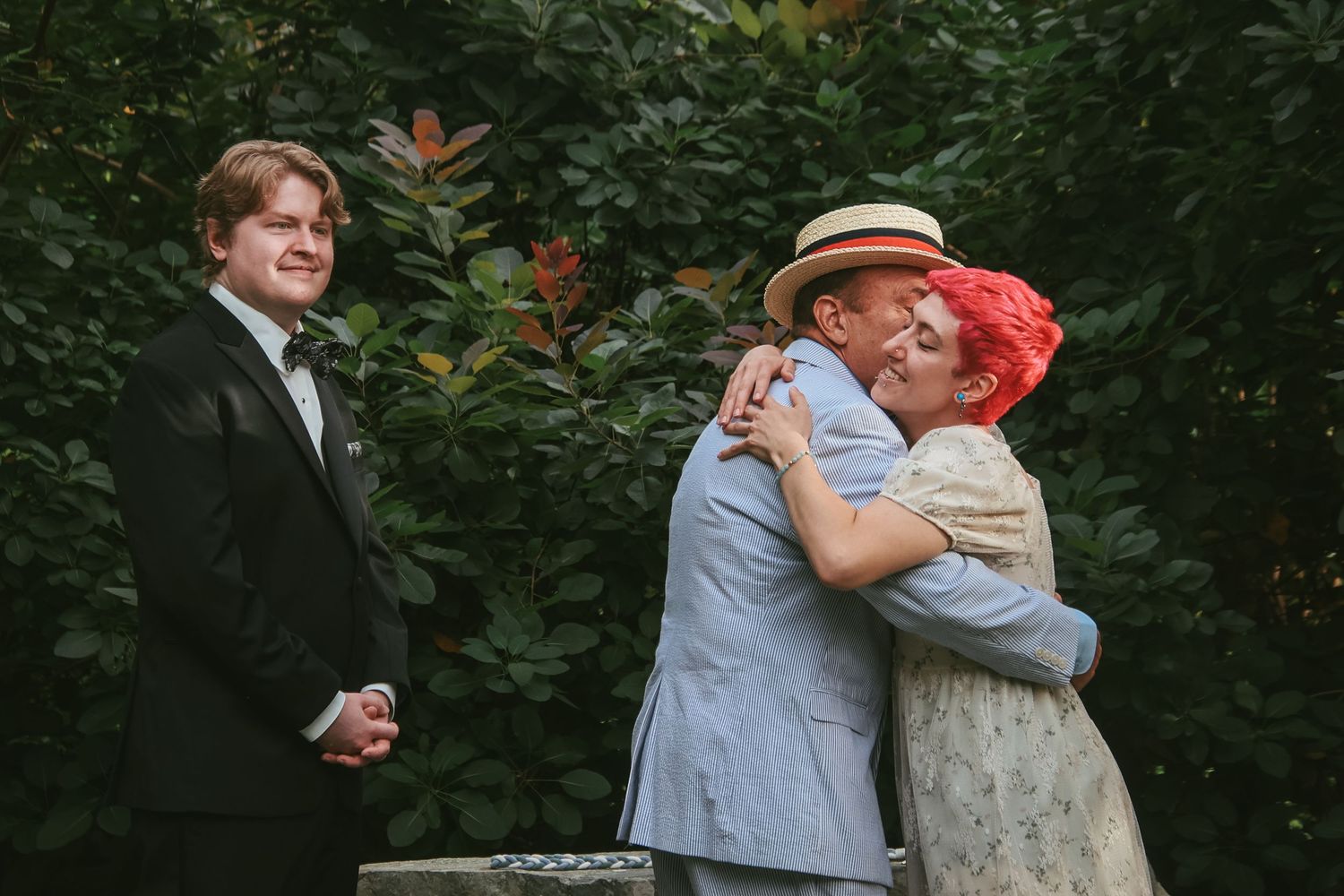 A tender embrace between two people in formal attire during an outdoor garden ceremony while another person looks on.