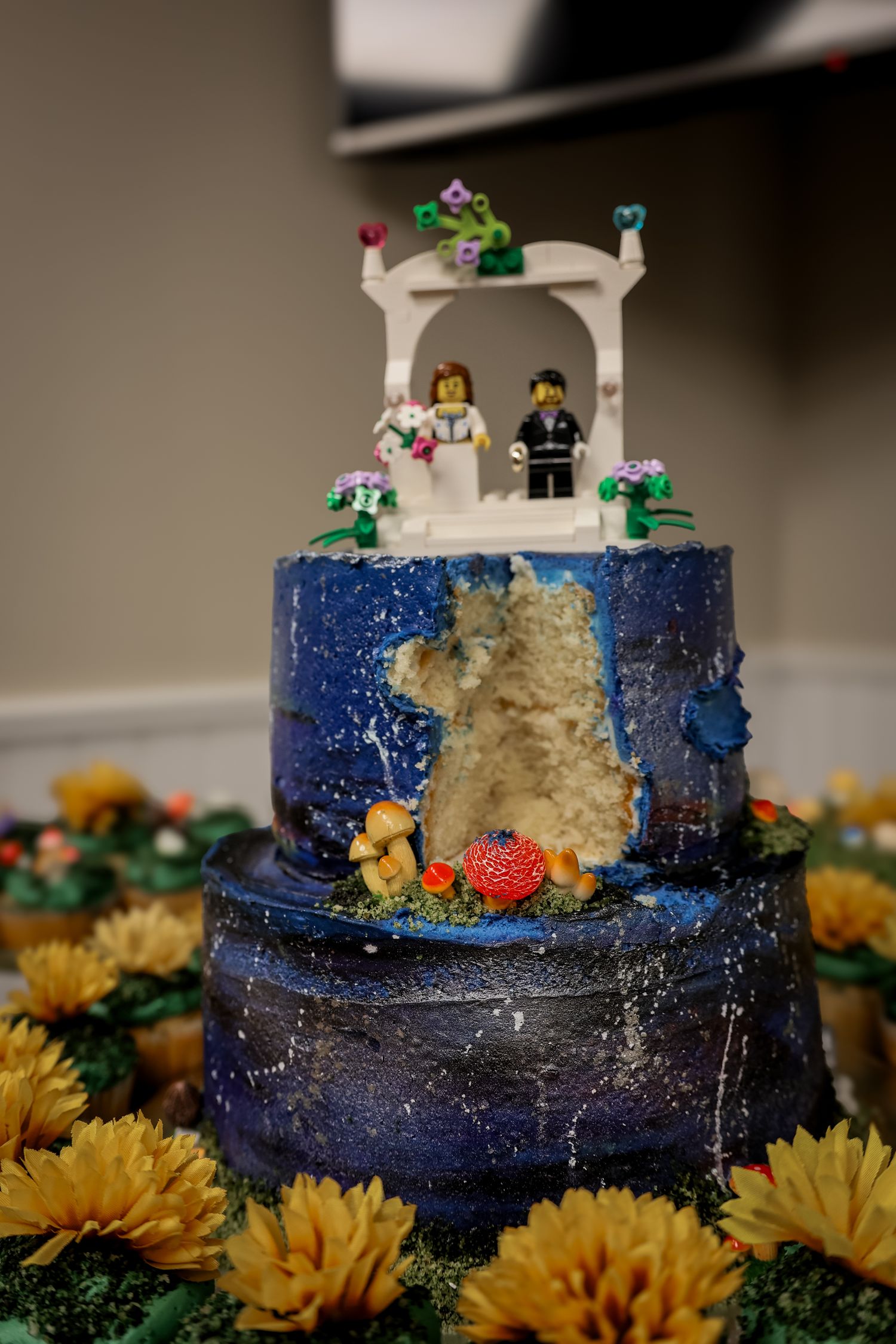 A navy blue wedding cake with gold accents decorated with yellow sunflowers and LEGO figurine toppers.