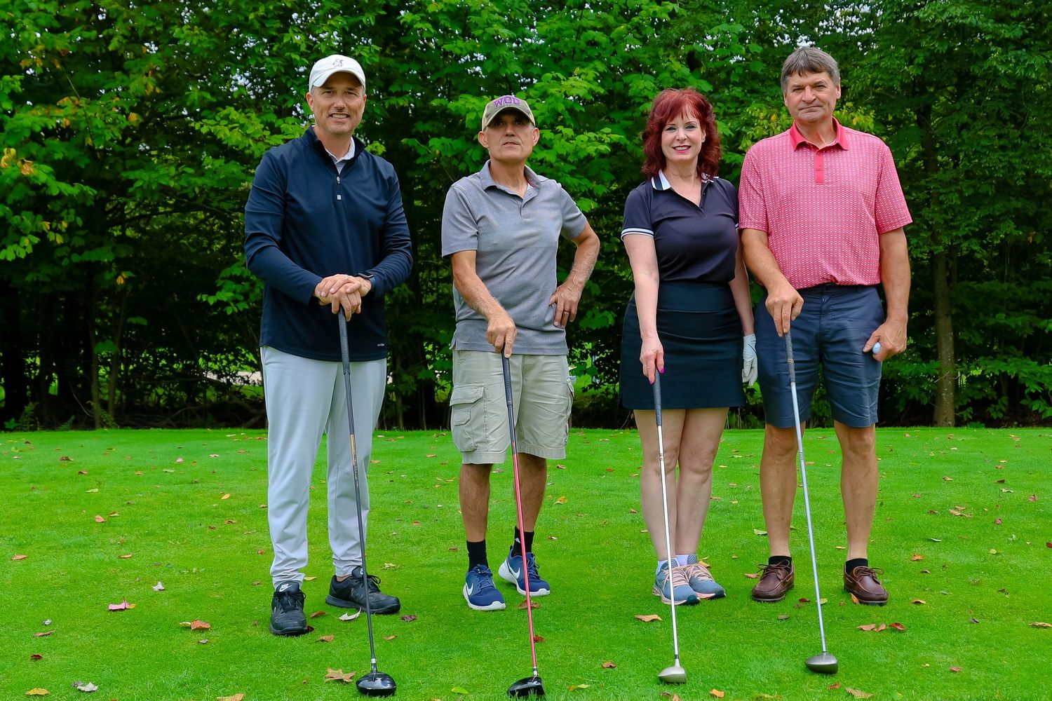 Four golfers with clubs pose together on a lush green golf course surrounded by trees.
